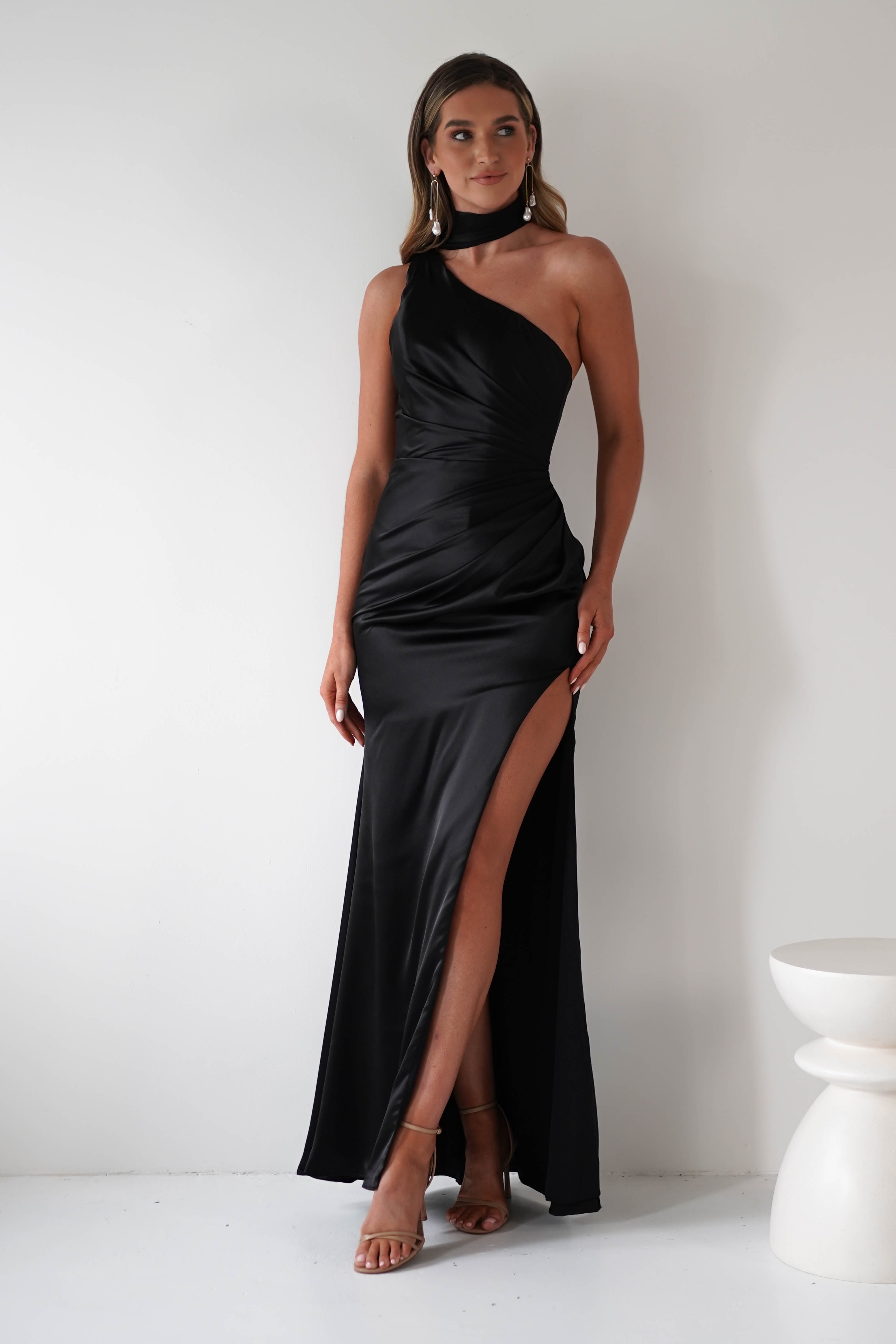 A woman stands against a white wall in a sleek, one-shoulder black satin gown with a gathered midriff and high thigh slit. She accessorizes with long earrings, strappy heels, and poses confidently with one leg slightly forward.