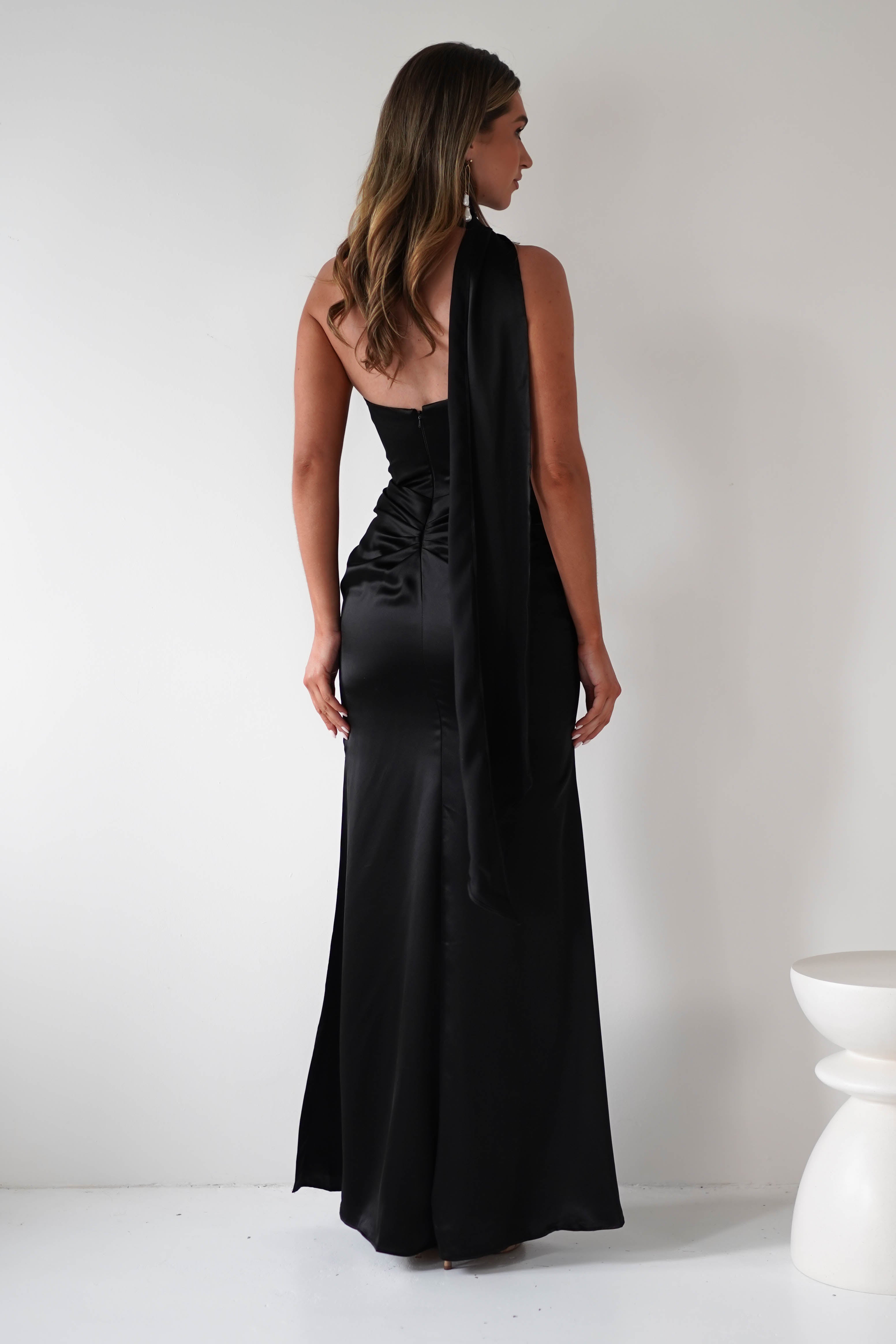 A woman with long, wavy hair stands facing away, wearing a floor-length, sleeveless black satin gown with an asymmetrical design and gathered midriff, in a minimalist white room.