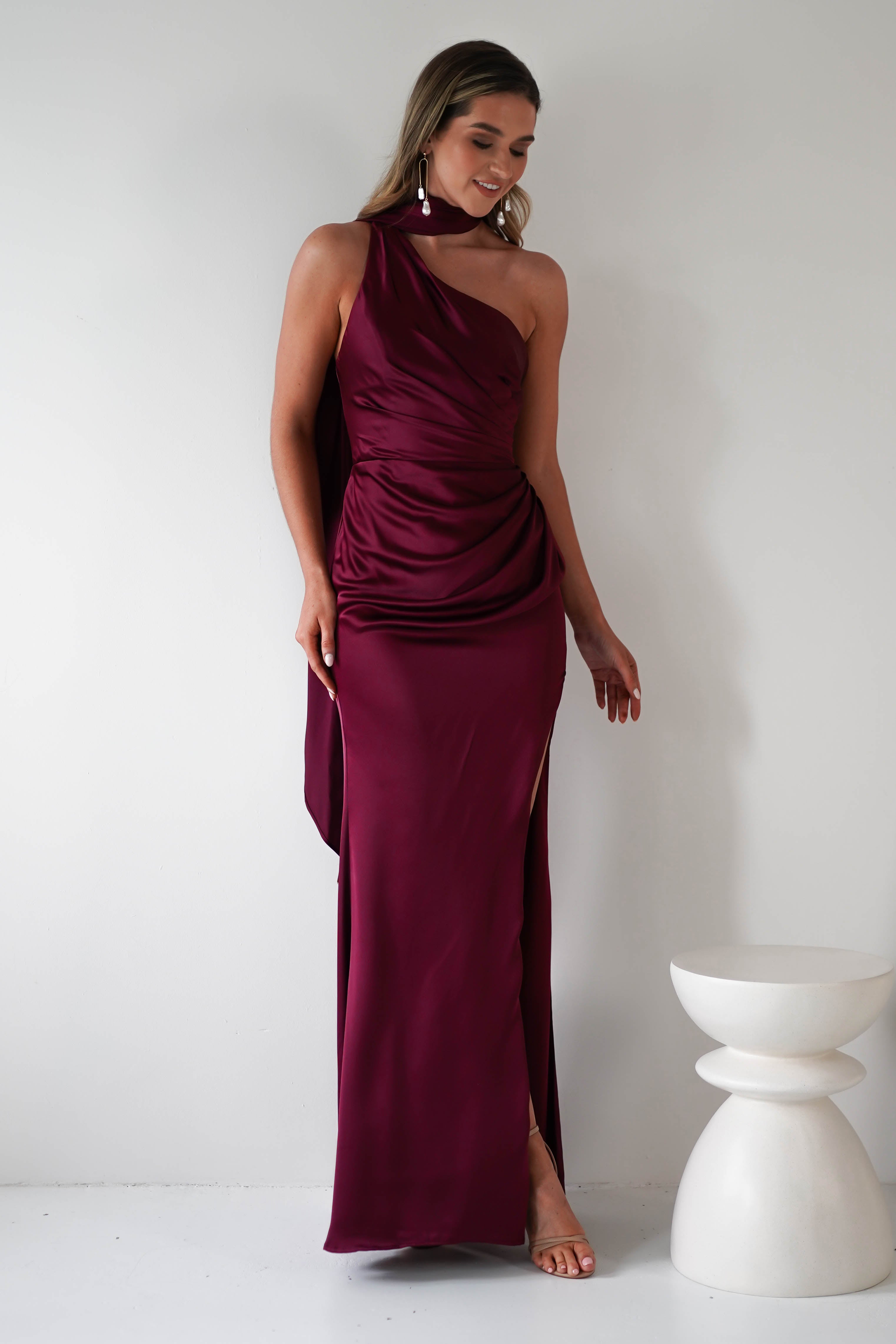 Woman in a sleeveless, burgundy satin dress with an asymmetrical neckline and thigh-high slit stands near a small, white sculptural table. Her gathered midriff subtly enhances the elegant silhouette. Shes smiling and wearing dangling earrings, set against a plain white background.