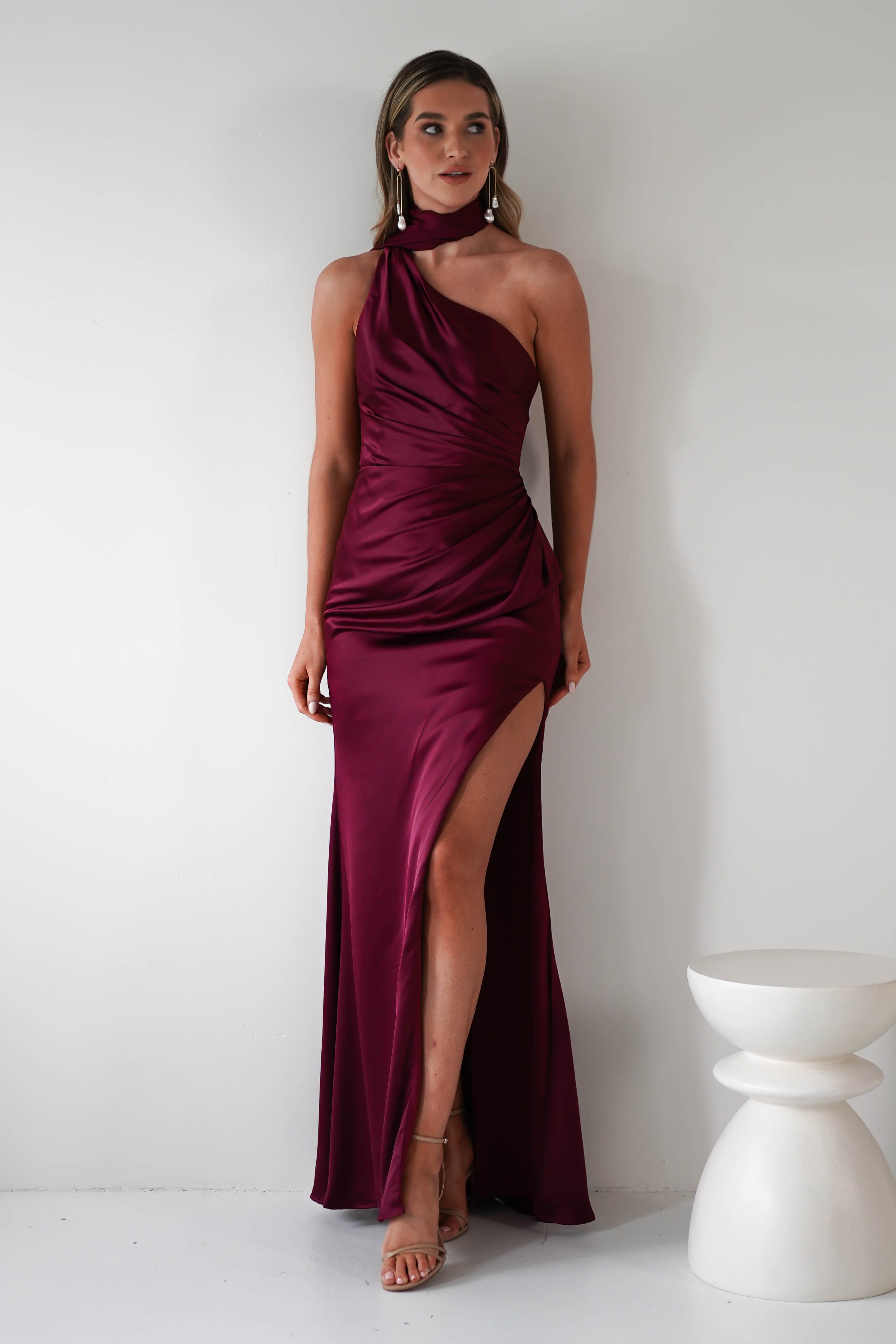 A woman stands against a white background, wearing a one-shoulder burgundy satin gown with a high slit and a gathered midriff. She has long hair and is accessorized with statement earrings. A white, modern stool is on her side.