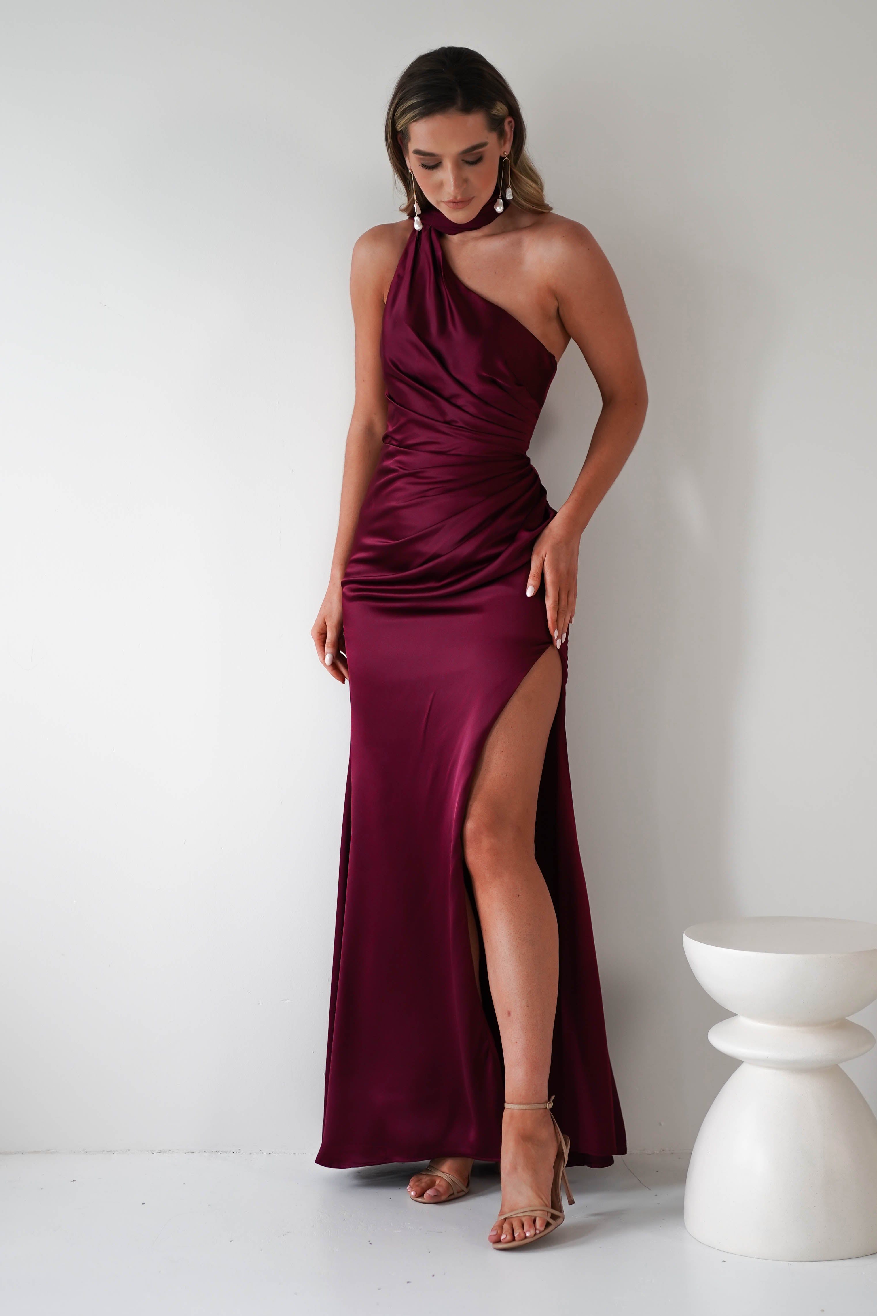 A person stands against a white wall in a burgundy, one-shoulder polyester satin gown with a gathered midriff and high slit. They have long hair, wearing dangling earrings and high-heeled sandals. A small white table is next to them.