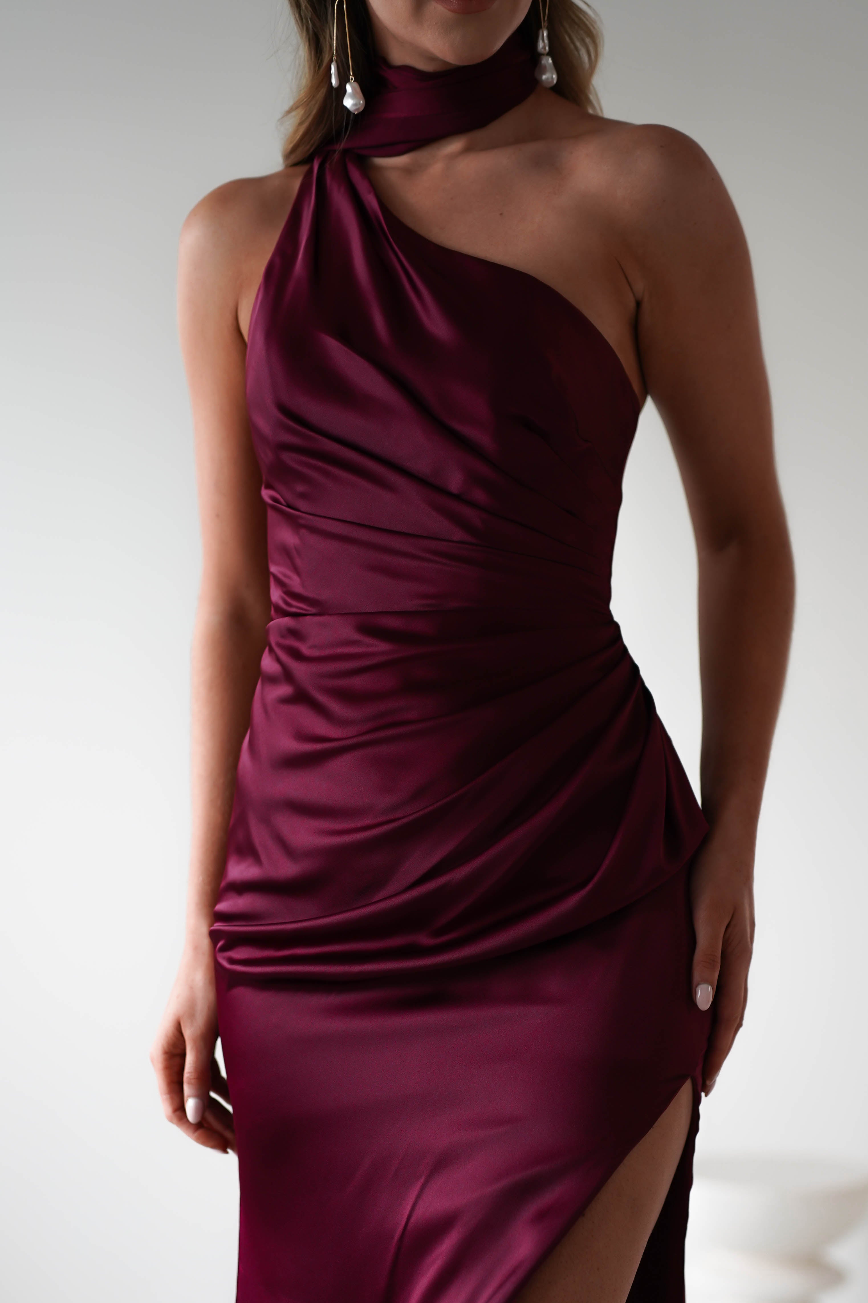 A person wearing a maroon, satin dress with gathered midriff detailing and a side slit. The neutral background enhances the elegant and sophisticated look of the polyester outfit, perfectly complemented by a stylish neck scarf.