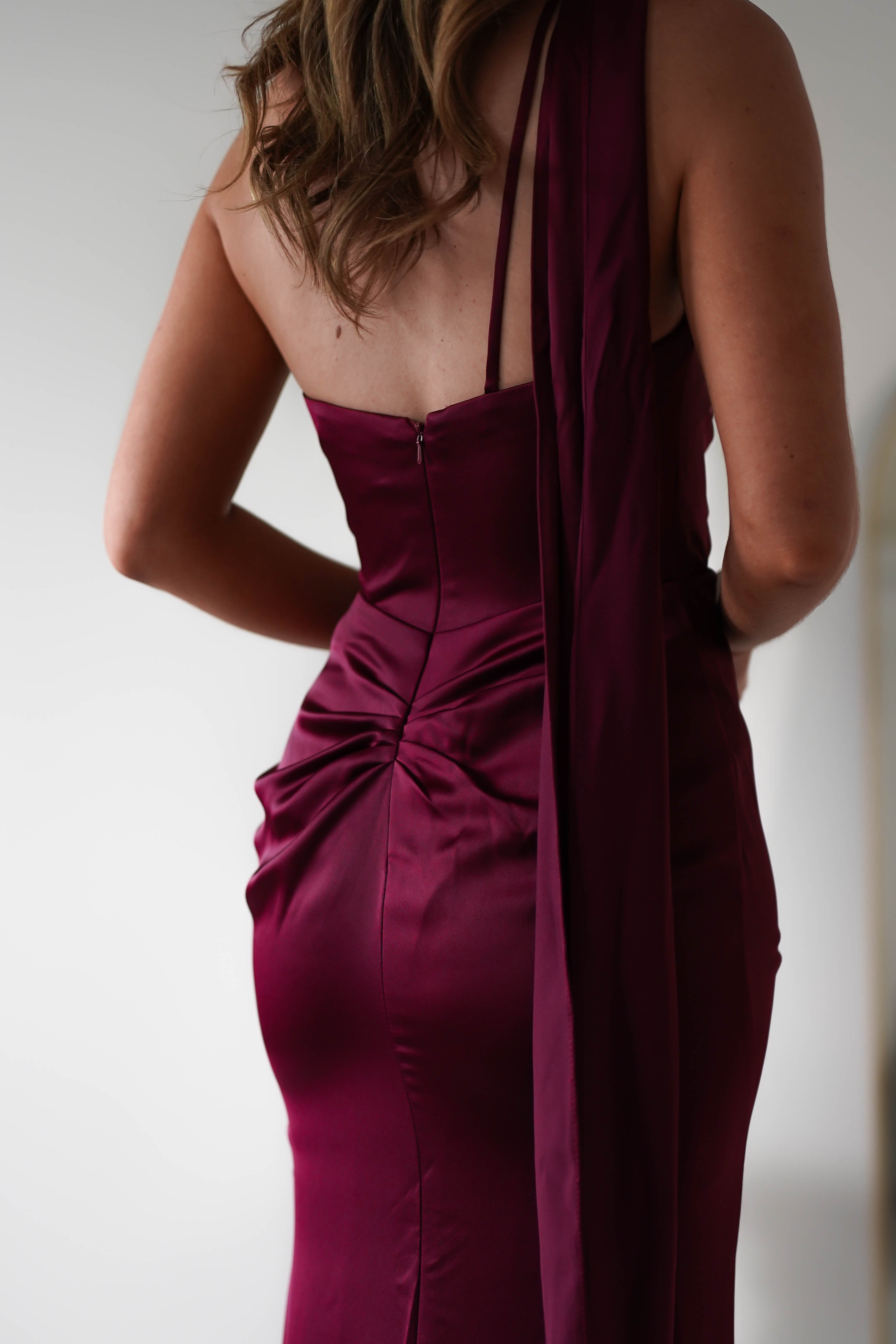 A person is wearing an elegant, sleeveless burgundy dress crafted from polyester, shown from the back. The dress features a fitted bodice, zip closure, and gathered fabric detailing. A chic neck scarf complements the look as their long, wavy hair cascades down their back.