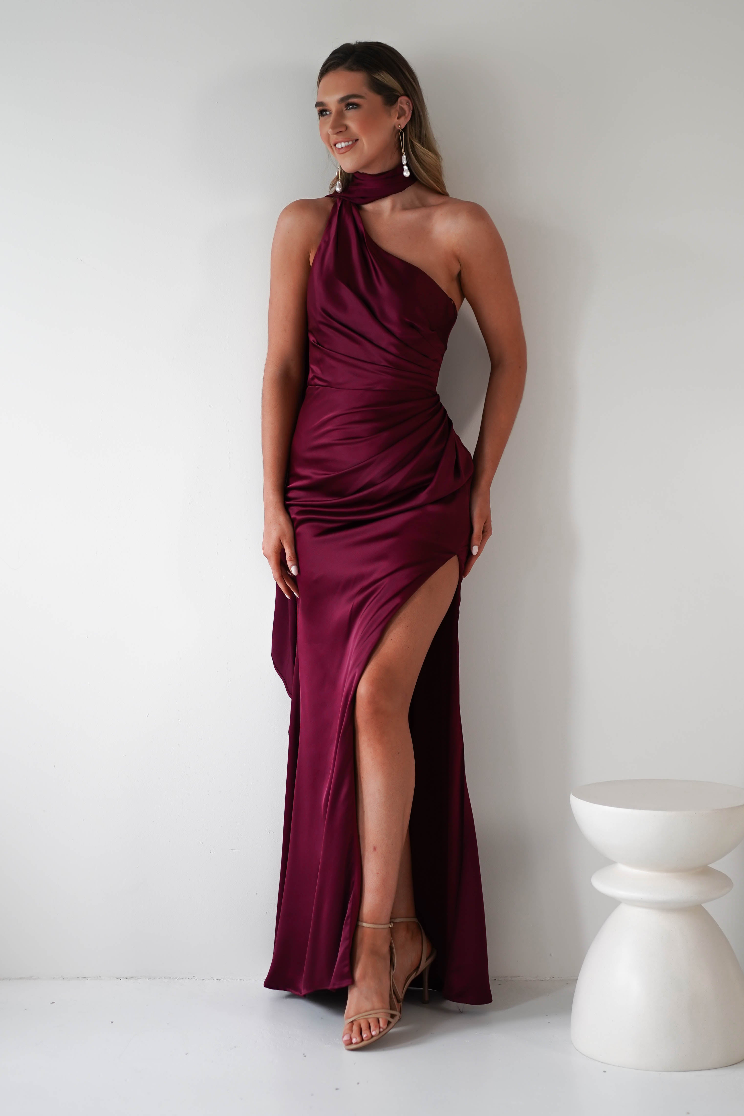 A woman in a burgundy, one-shoulder satin gown with a thigh-high slit and gathered midriff leans against a white wall. She has long hair, wearing earrings and sandals. A small white stool is beside her.