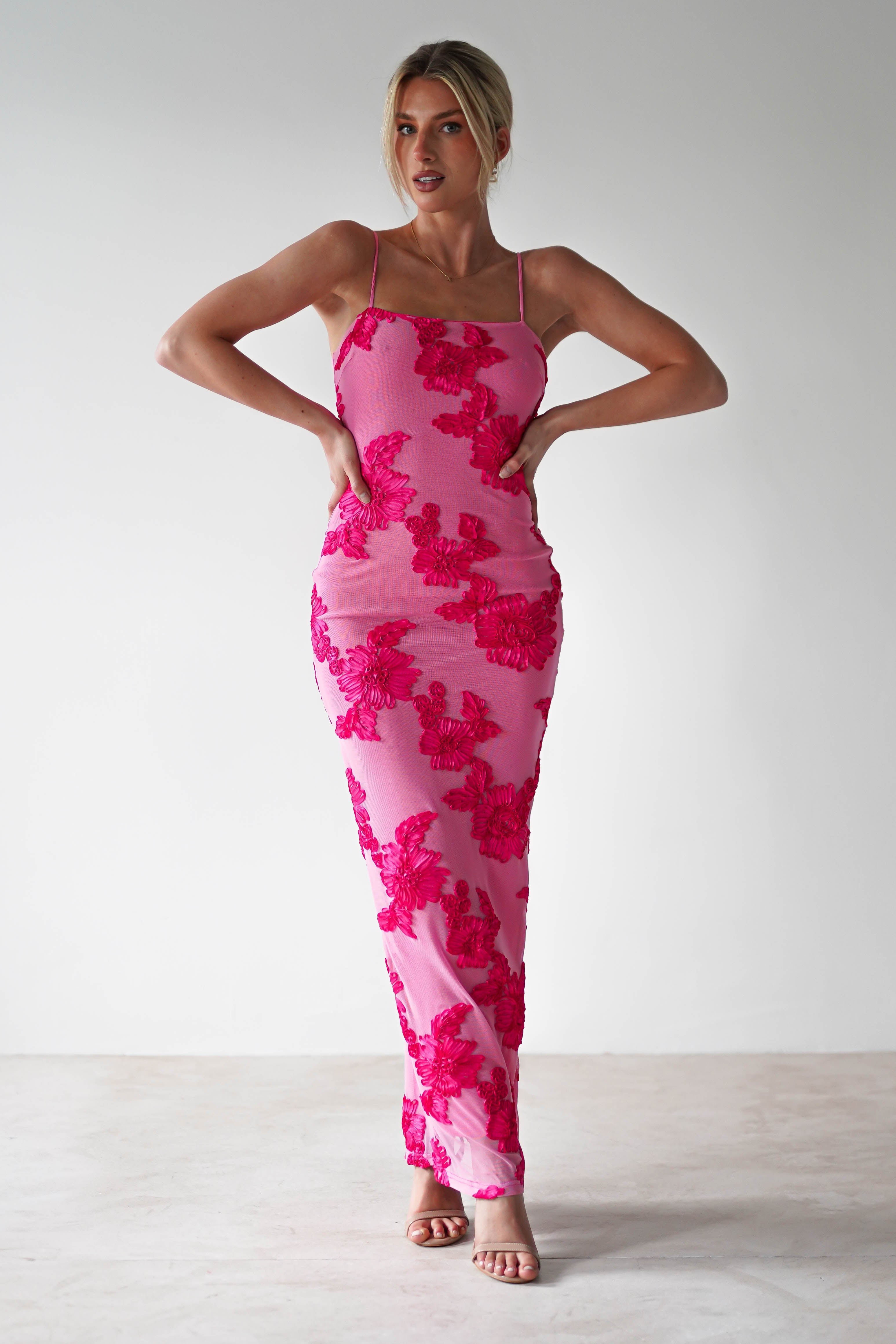 Woman in a pink, form-fitting midaxi dress with red floral patterns, standing with hands on hips. The dress is perfect for wedding guest attire, paired elegantly with heeled sandals. She has blonde hair styled up, set against a plain background.