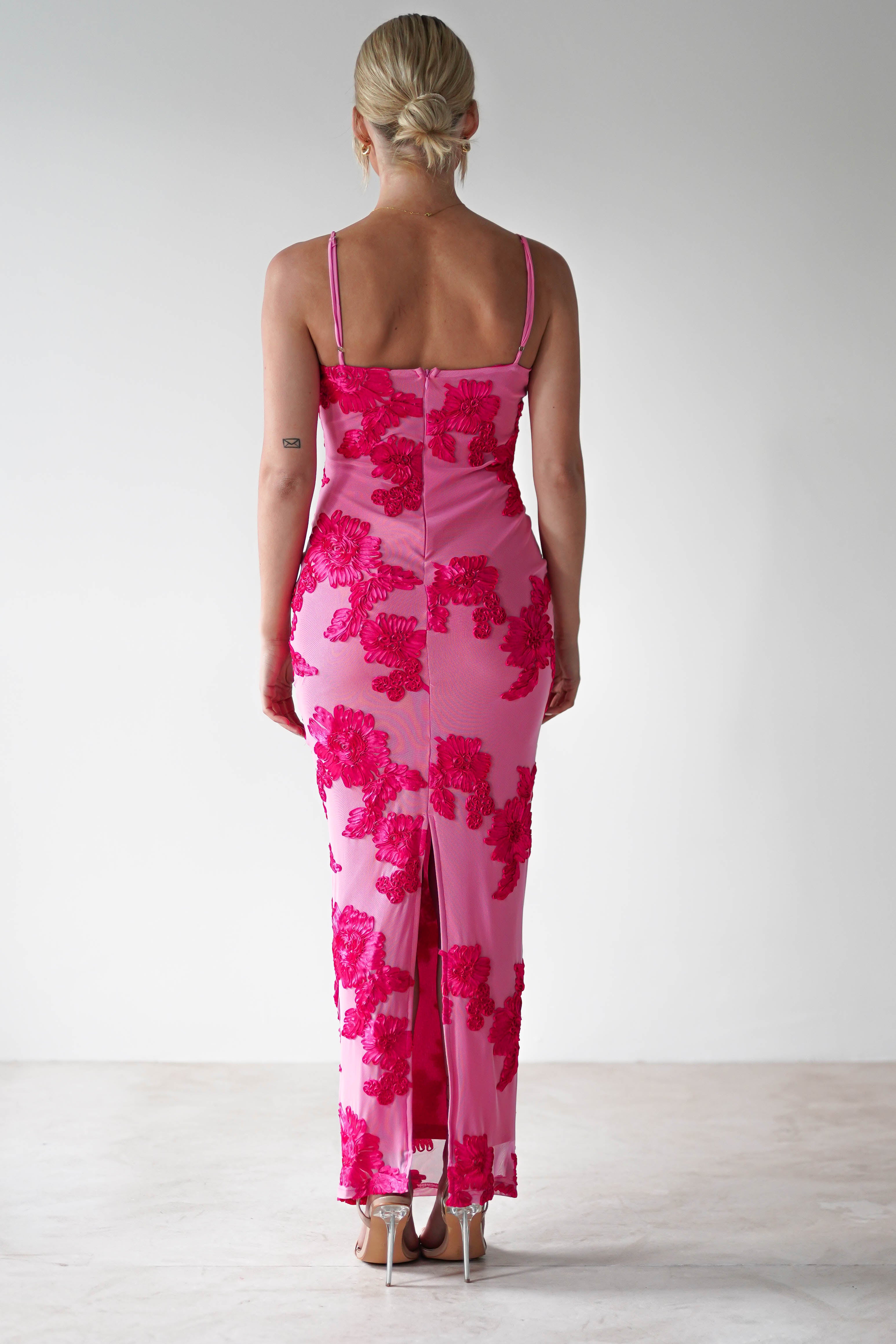 A person with blonde hair is facing away, wearing a form-fitting, stretchy midaxi dress in pink with bold floral patterns. The dress features thin straps and a back slit, paired elegantly with high heels, perfect for wedding guest attire against a plain white background.