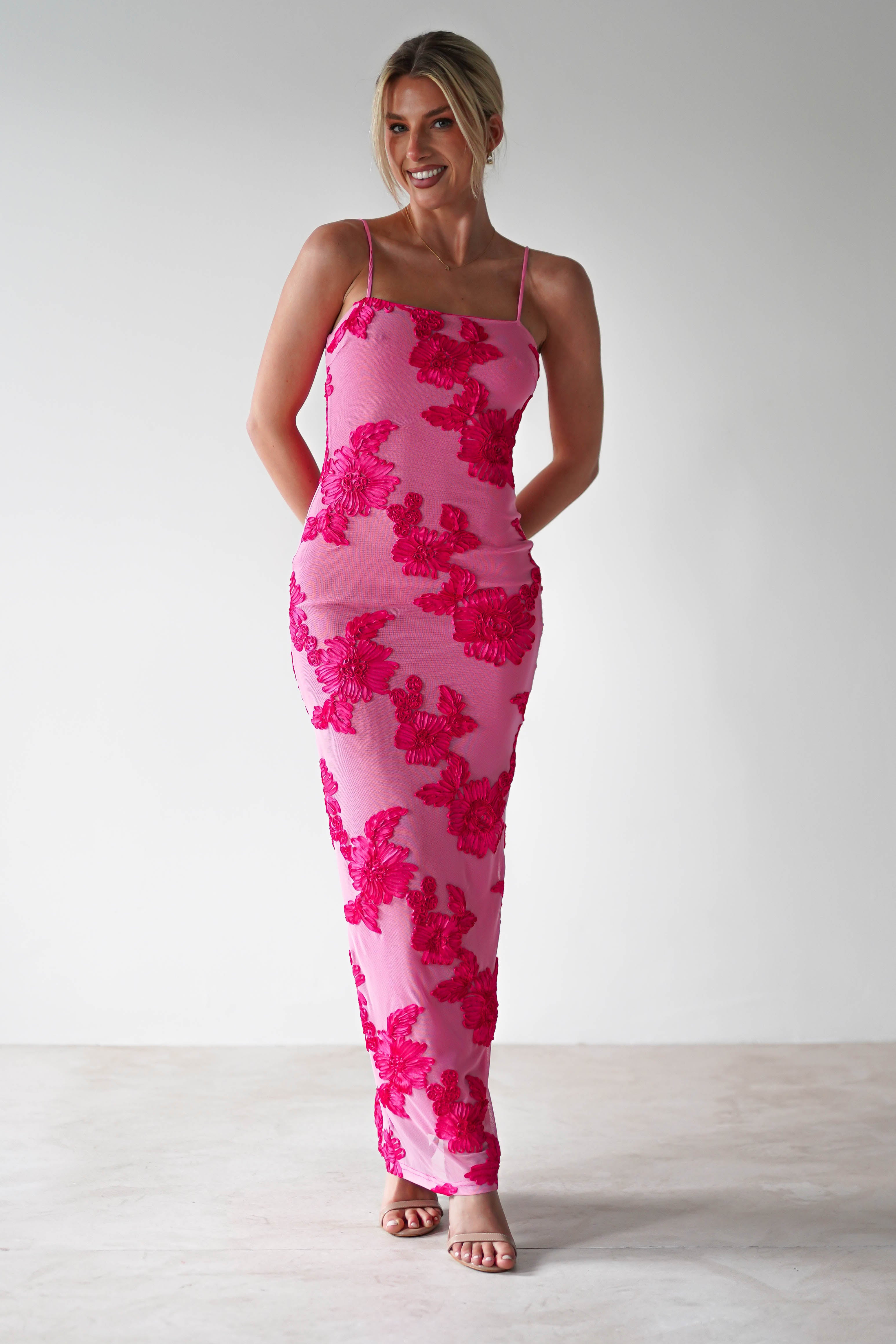 A person stands in a pink midaxi dress adorned with red floral patterns, perfect for wedding guest attire. They have a cheerful expression, with their hands behind their back. Wearing strappy sandals, they gracefully pose against a simple white background.