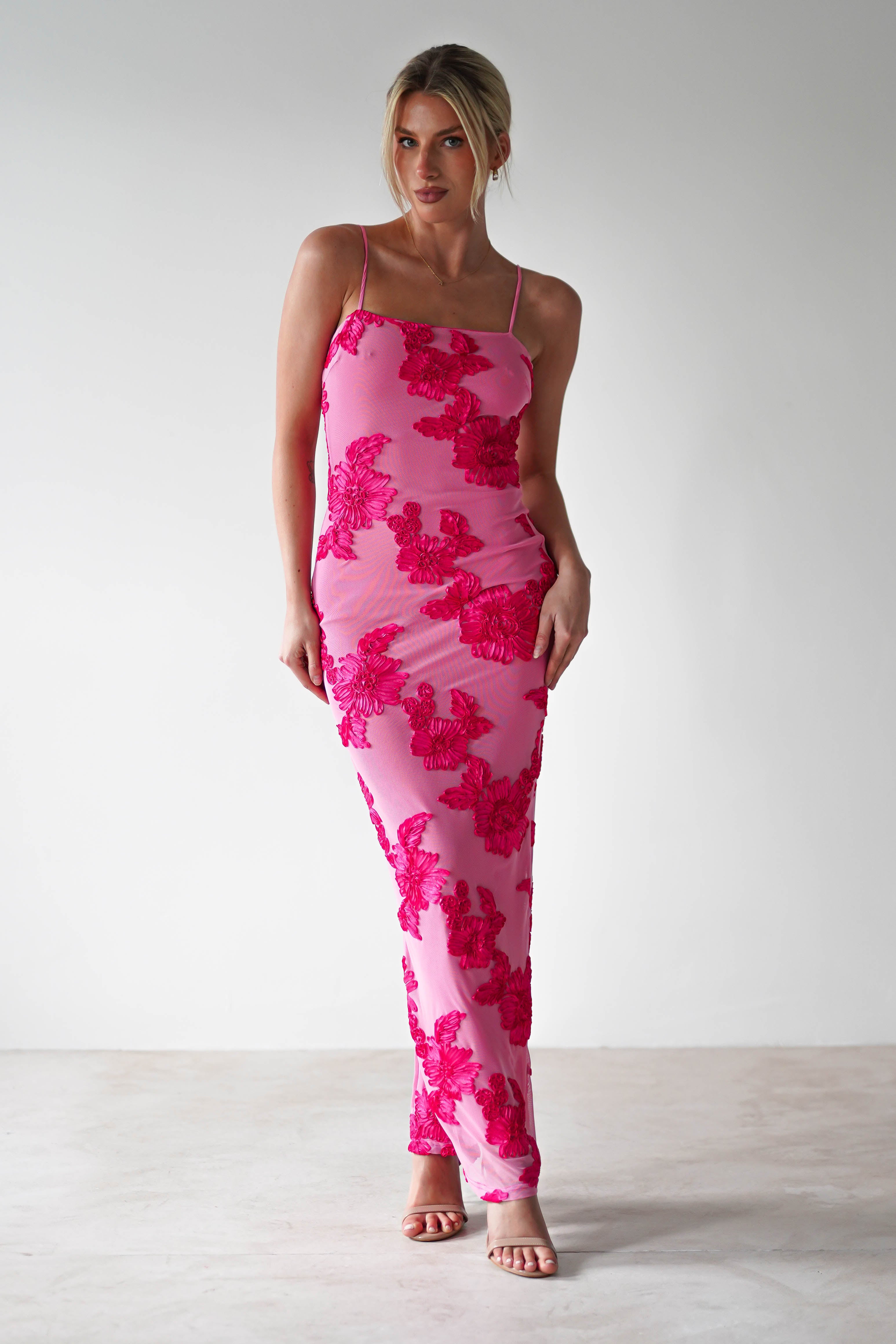 A person poses gracefully in front of a plain white background, wearing a fitted pink midaxi dress with bold red floral patterns. Their light-colored hair is styled up, and strappy pink heels complete their chic wedding guest attire.