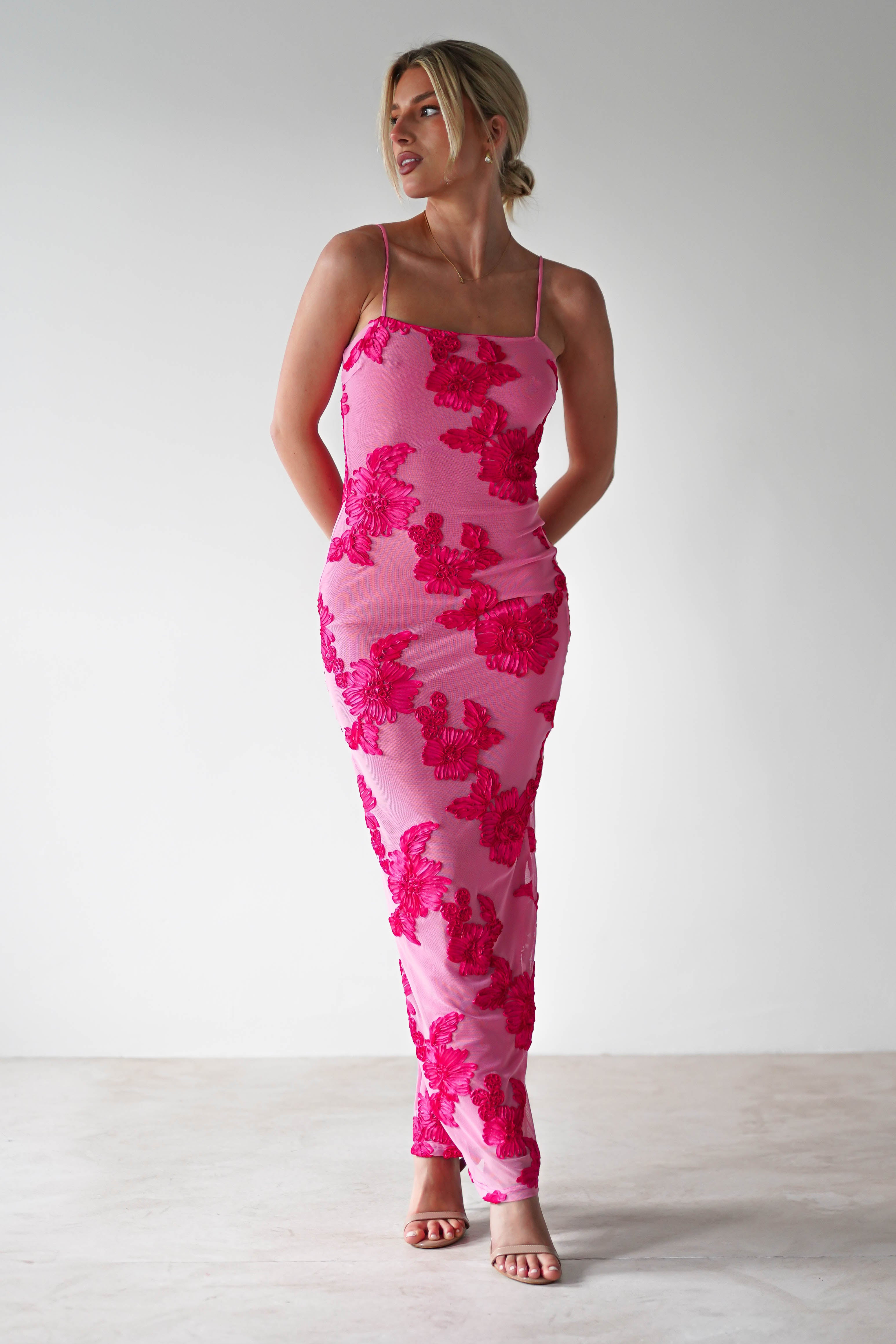 A person wearing a form-fitting midaxi pink dress with floral patterns stands against a plain white background, looking to the right. They have blonde hair styled up and are wearing strappy pink heels—perfect wedding guest attire.