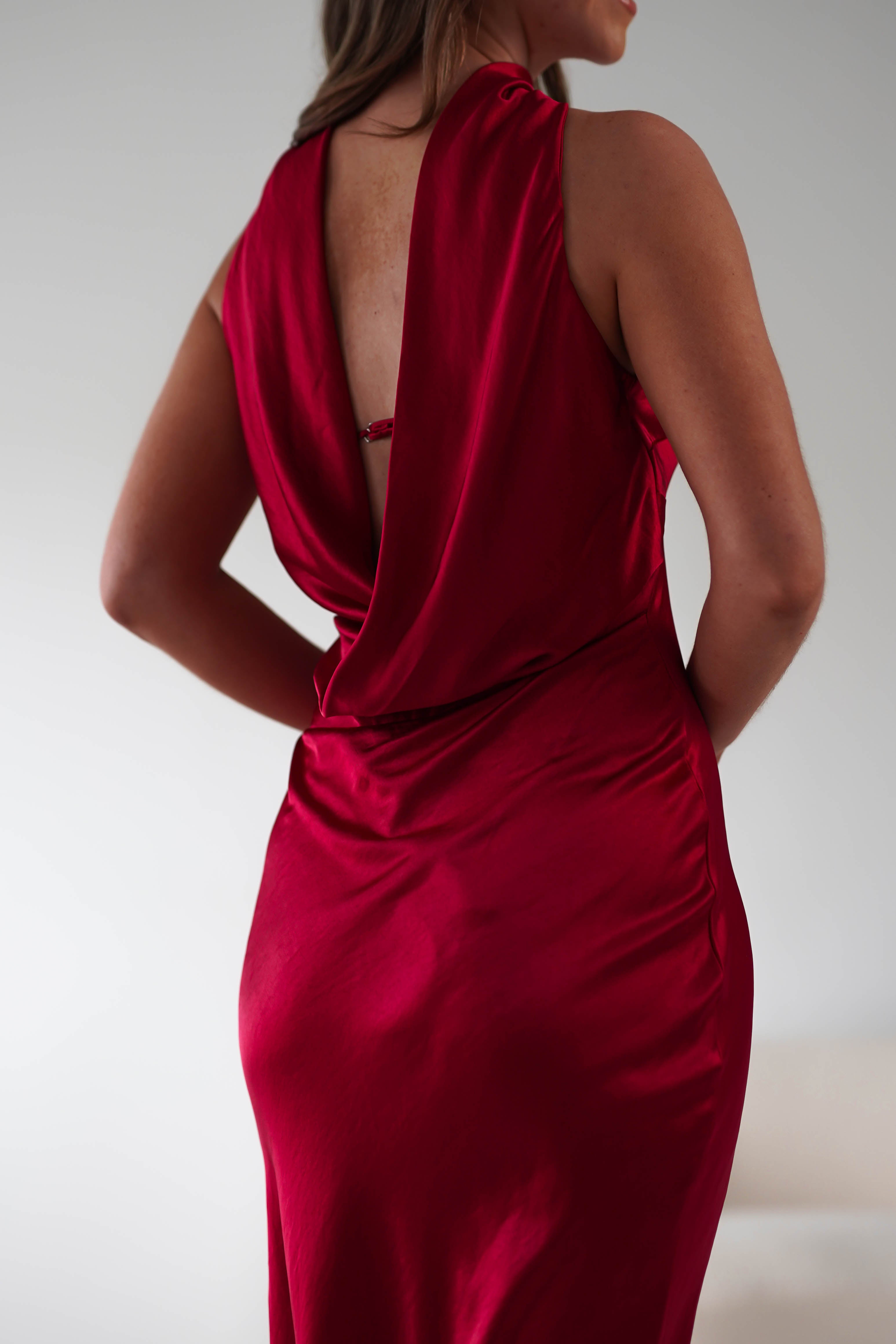 A woman wearing a backless red satin maxi dress stands facing away, her hands gracefully resting on her hips. Perfect for special occasions, the elegant drape of the dress highlights its smooth texture against a simple, neutral background.