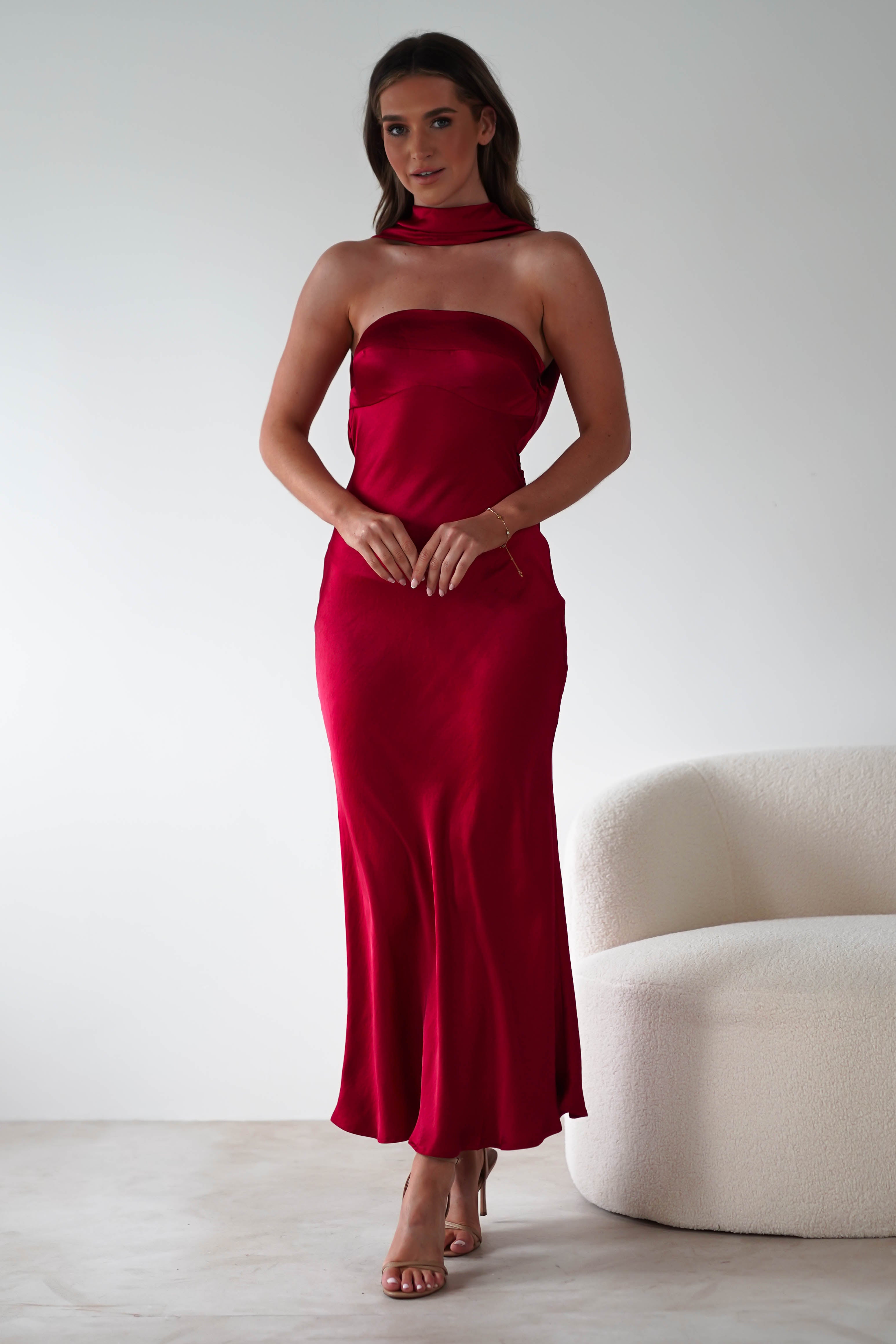 A woman in a strapless satin maxi dress stands against a white background near a soft white chair. Perfect for special occasions, her minimal jewelry complements the floor-length red dress, with long brown hair cascading over her shoulders and hands elegantly clasped in front of her.