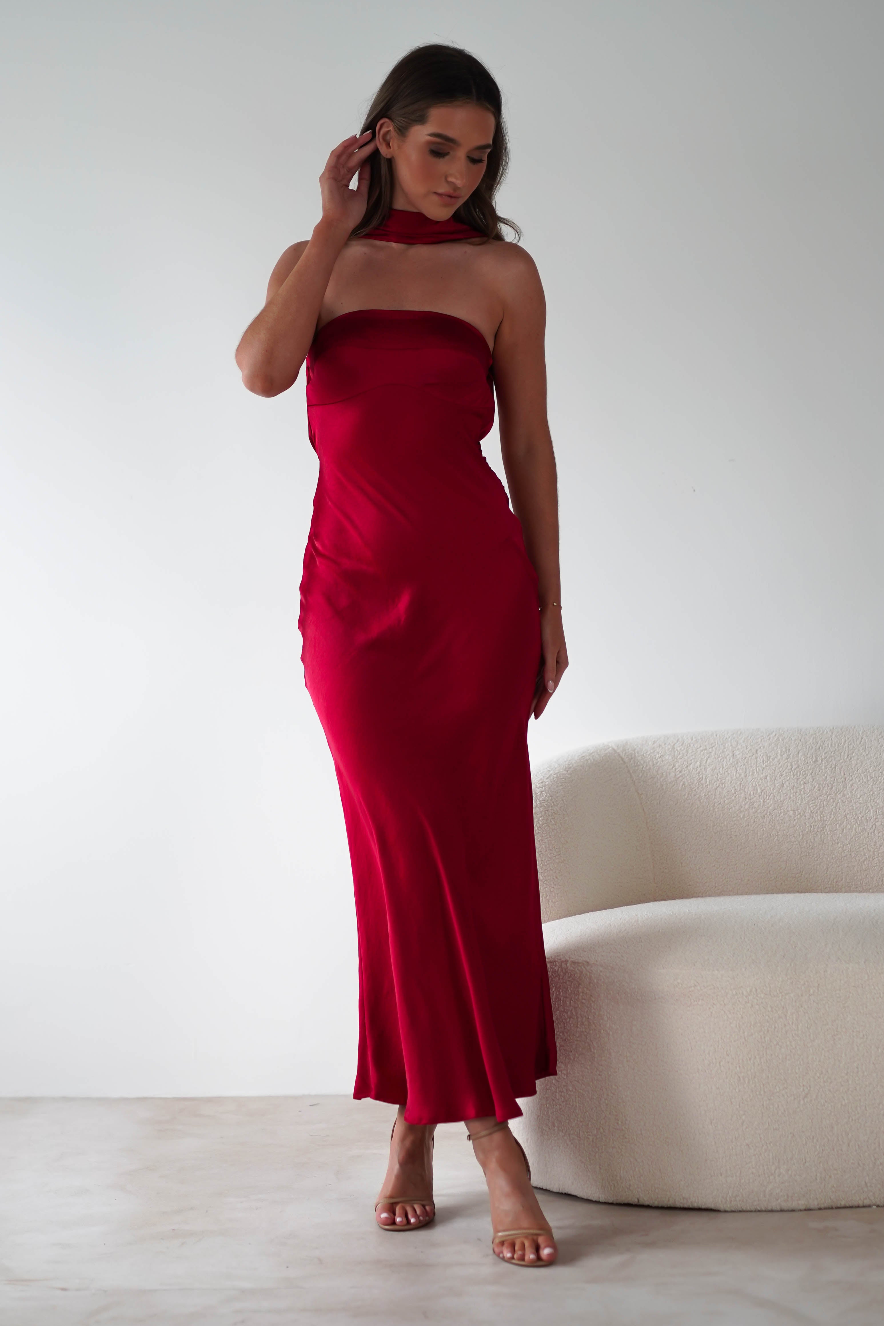 A woman in a satin maxi dress stands near a cream-colored sofa against a plain white background. Perfect for special occasions, the long, strapless red gown complements her flowing hair as she gracefully touches it with one hand and rests the other on her hip.