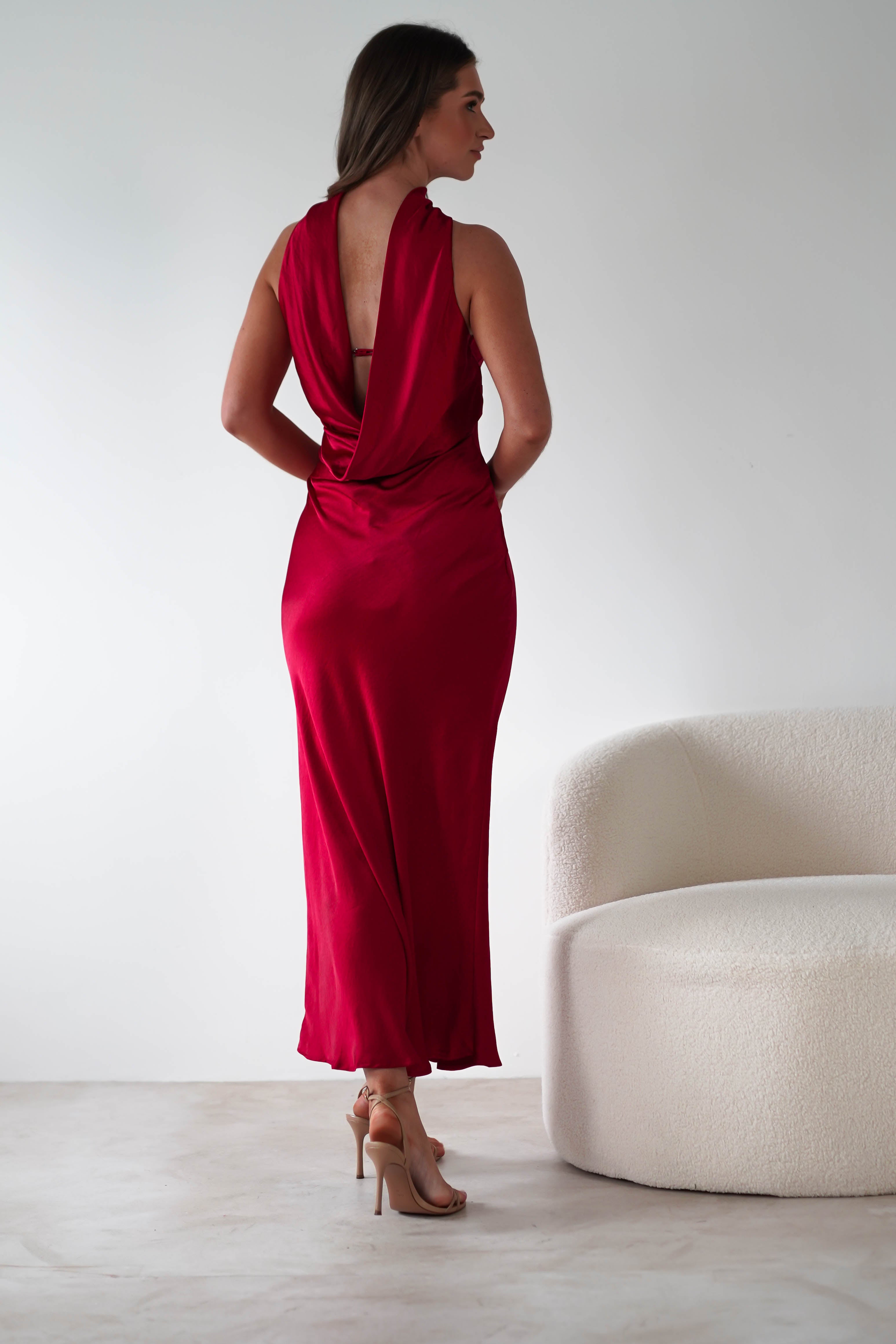A person stands with their back to the camera, wearing a stunning floor-length, sleeveless satin maxi dress in red, perfect for special occasions. They are next to a white, modern, curved armchair in a minimalist setting.