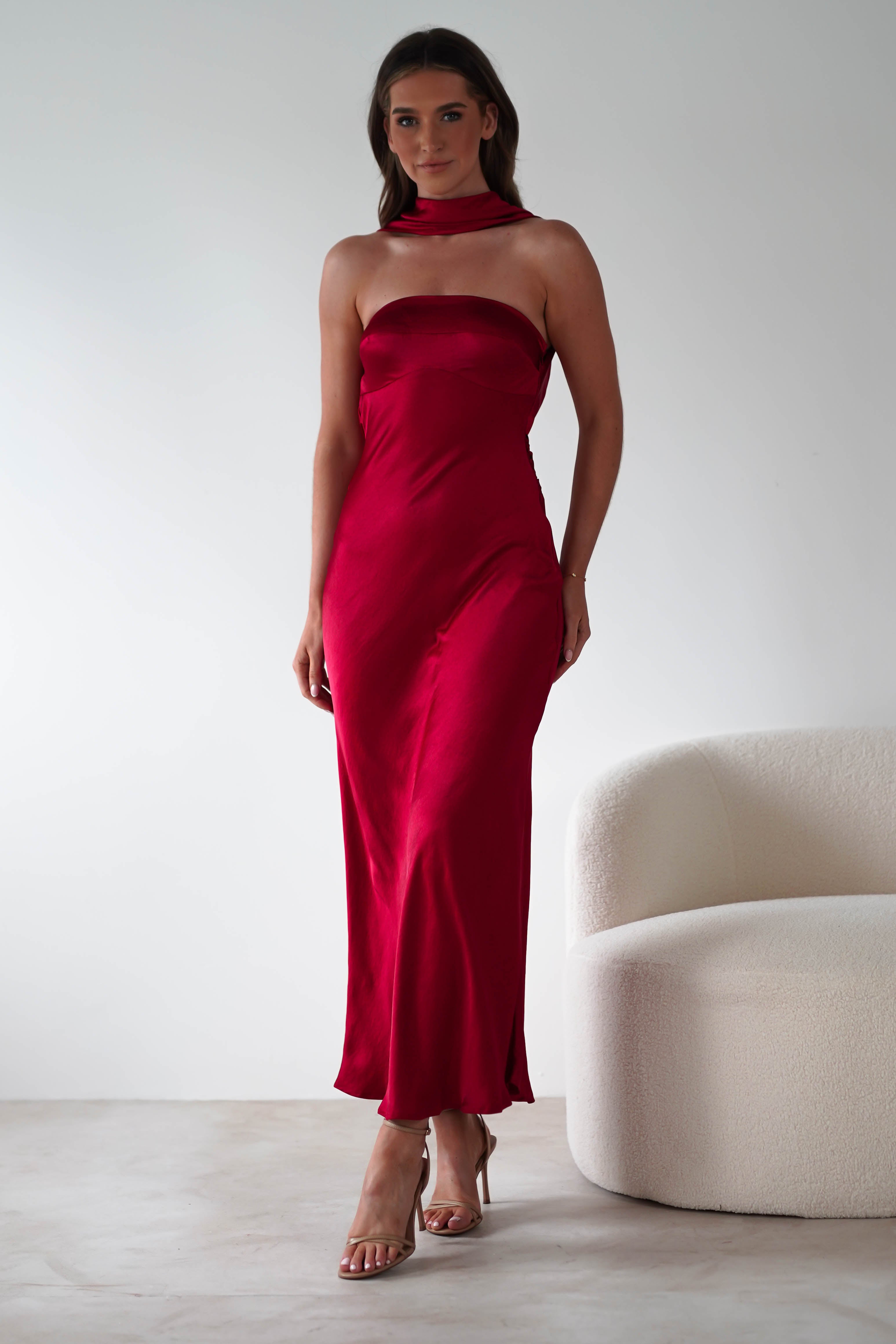 A woman in a strapless red satin maxi dress stands beside a cream-colored textured chair against a plain background, ready for special occasions or as an elegant wedding guest.
