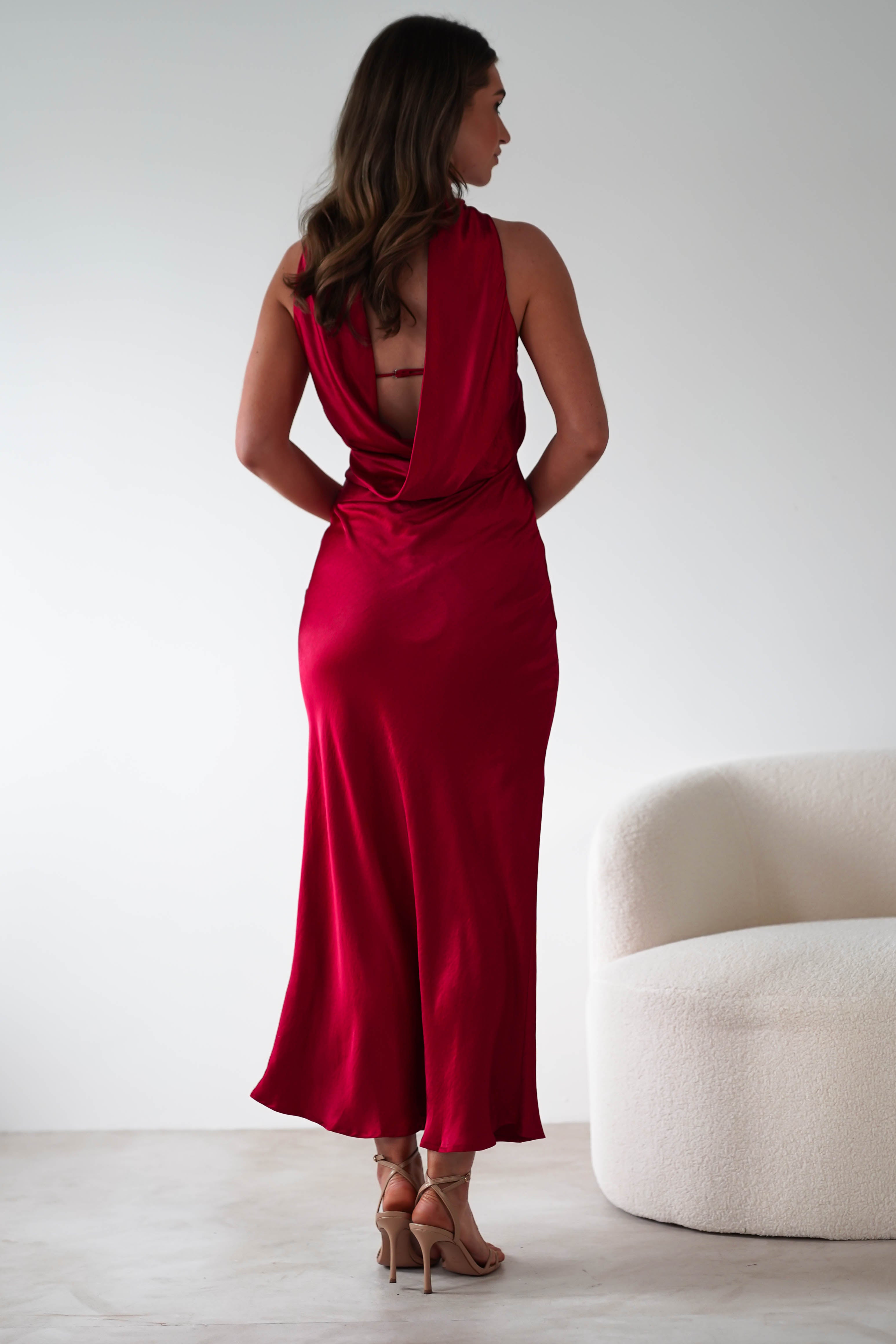 A woman in a flowing red satin maxi dress stands with her back to the camera in a minimalist room. Perfect for special occasions, the dress with an open back drapes elegantly. She wears beige high heels and stands near a modern white chair.
