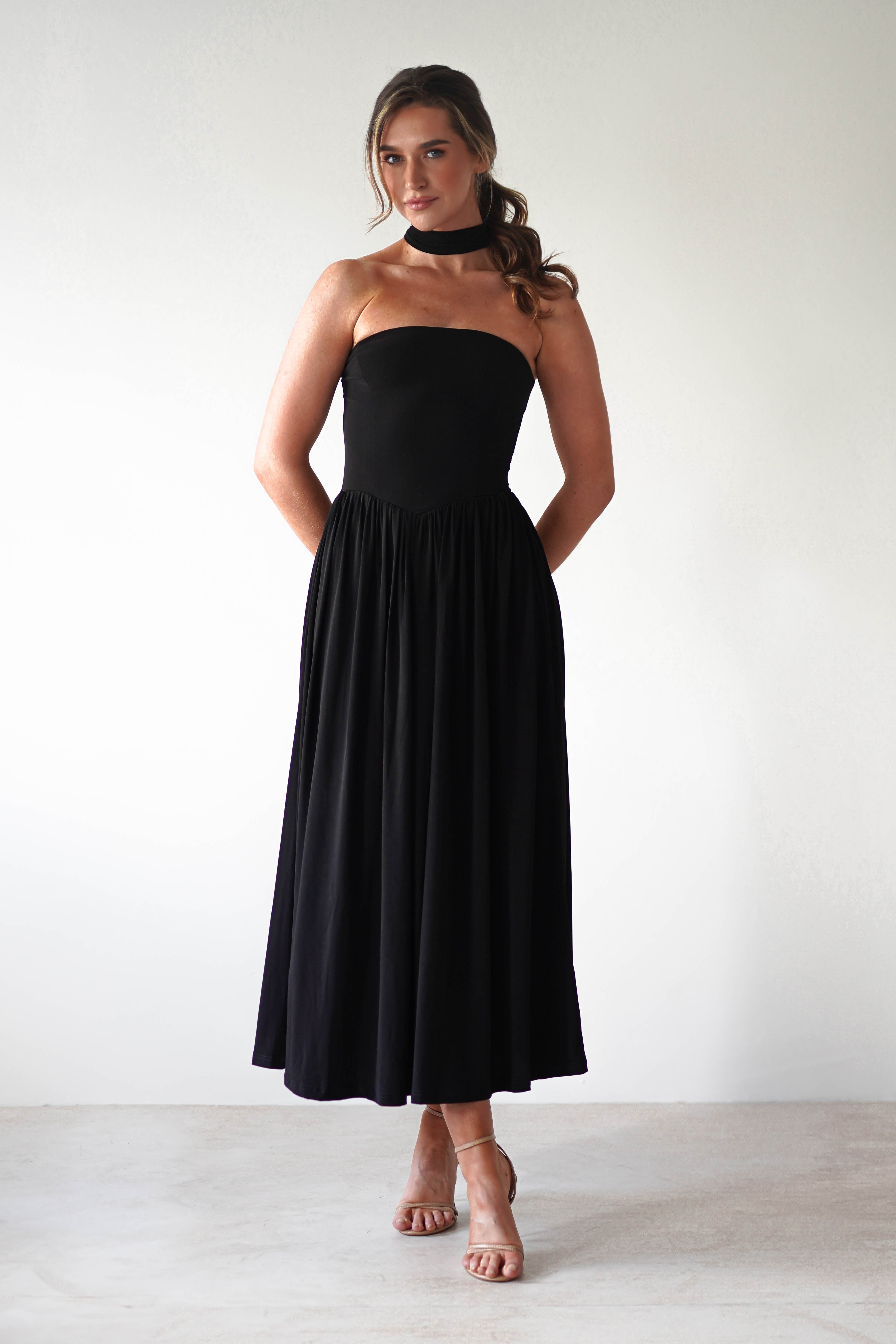 A woman stands against a plain light background, wearing a strapless black midi dress with a pleated skirt and stretchy fabric—perfect for a stylish wedding guest. She completes the look with a black choker, nude heels, and loose waves in her hair.