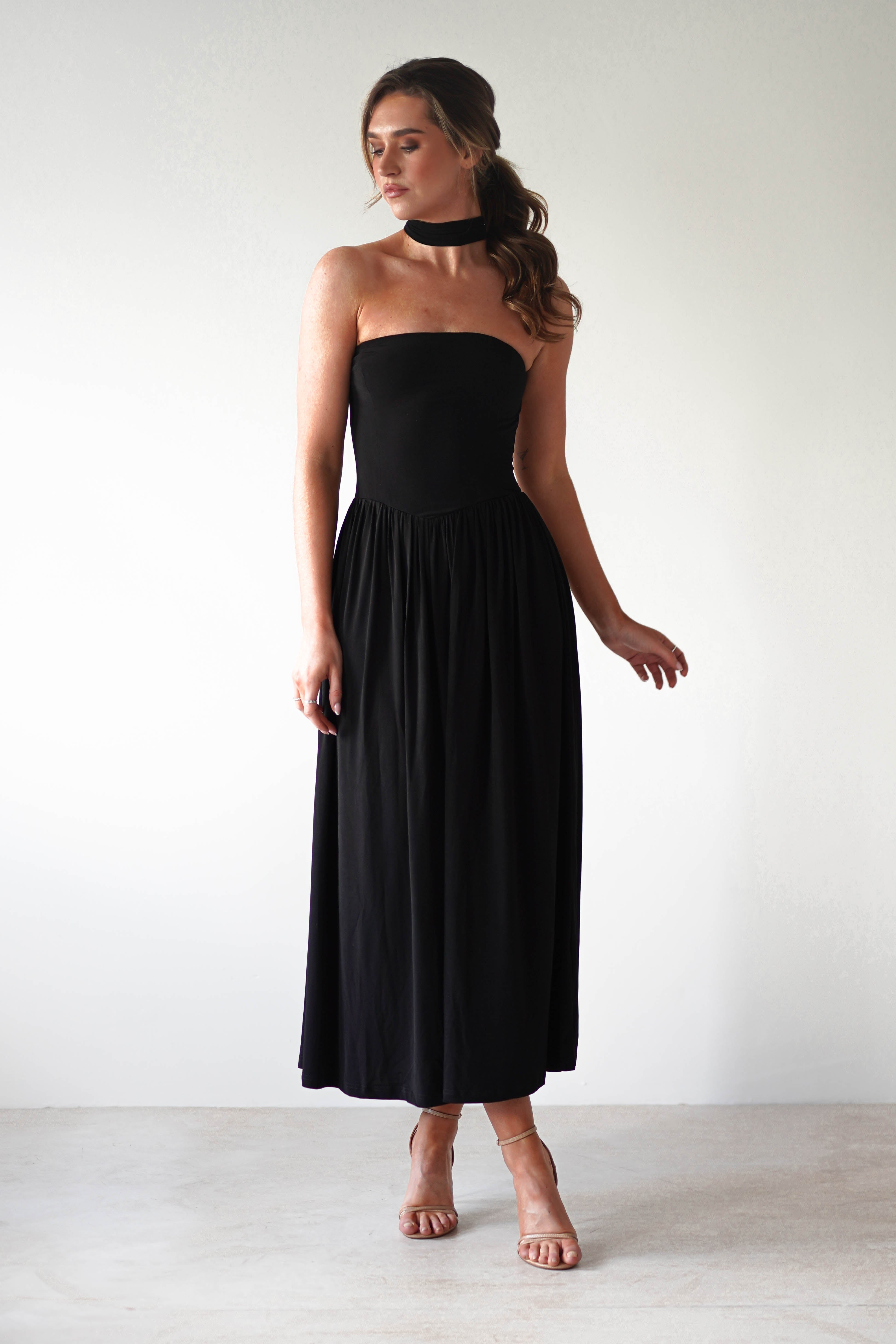 A woman wearing a strapless black dress made from stretchy fabric and nude heels stands against a plain white background. She has wavy hair styled half-up and accessorizes with a chic black choker necklace.