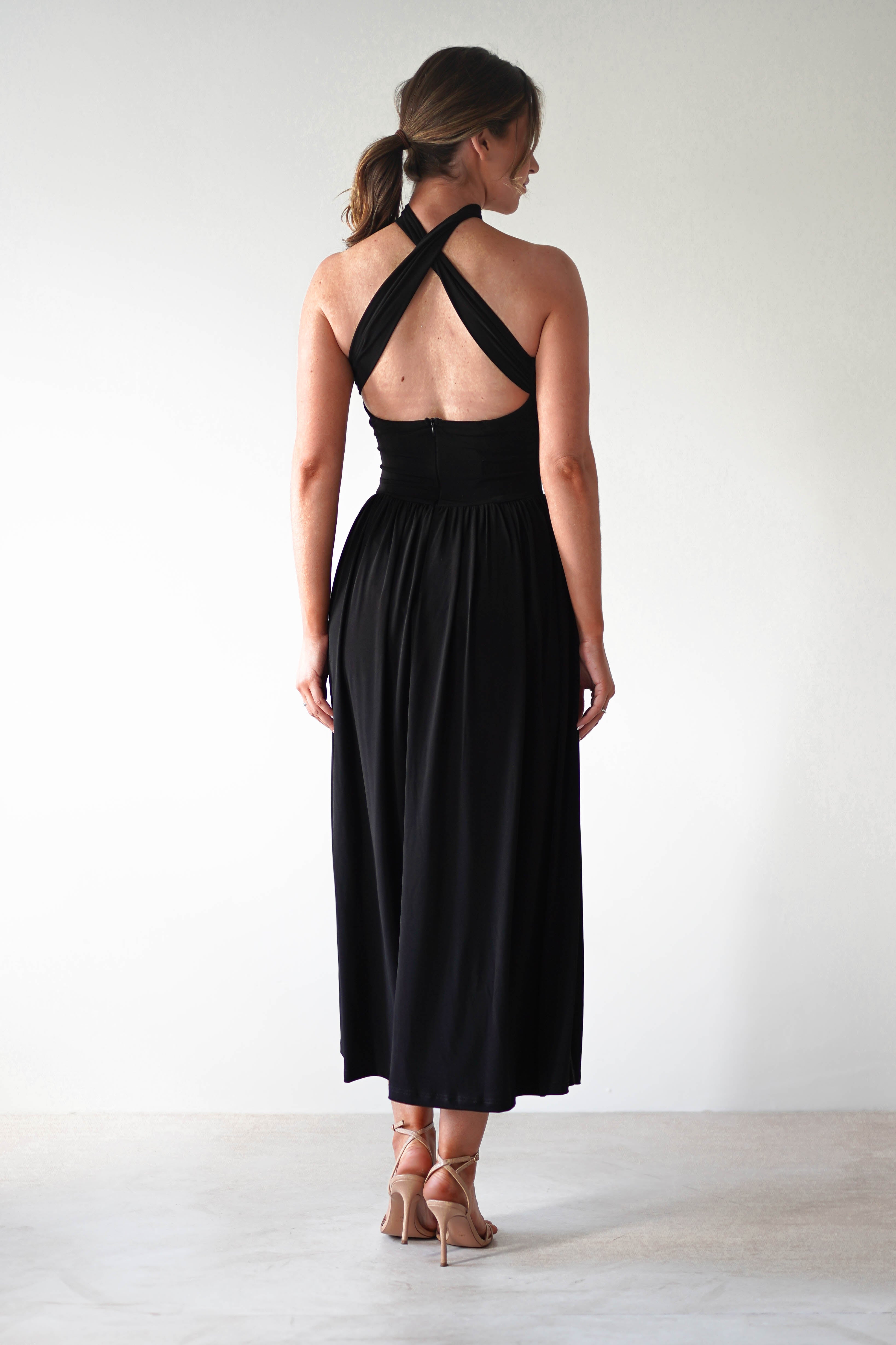 A woman stands facing away, wearing a sleeveless black midi dress with a crisscross halter neckline and a pleated skirt in stretchy fabric—perfect for a wedding guest. Her hair is tied back in a loose ponytail against a plain light background.