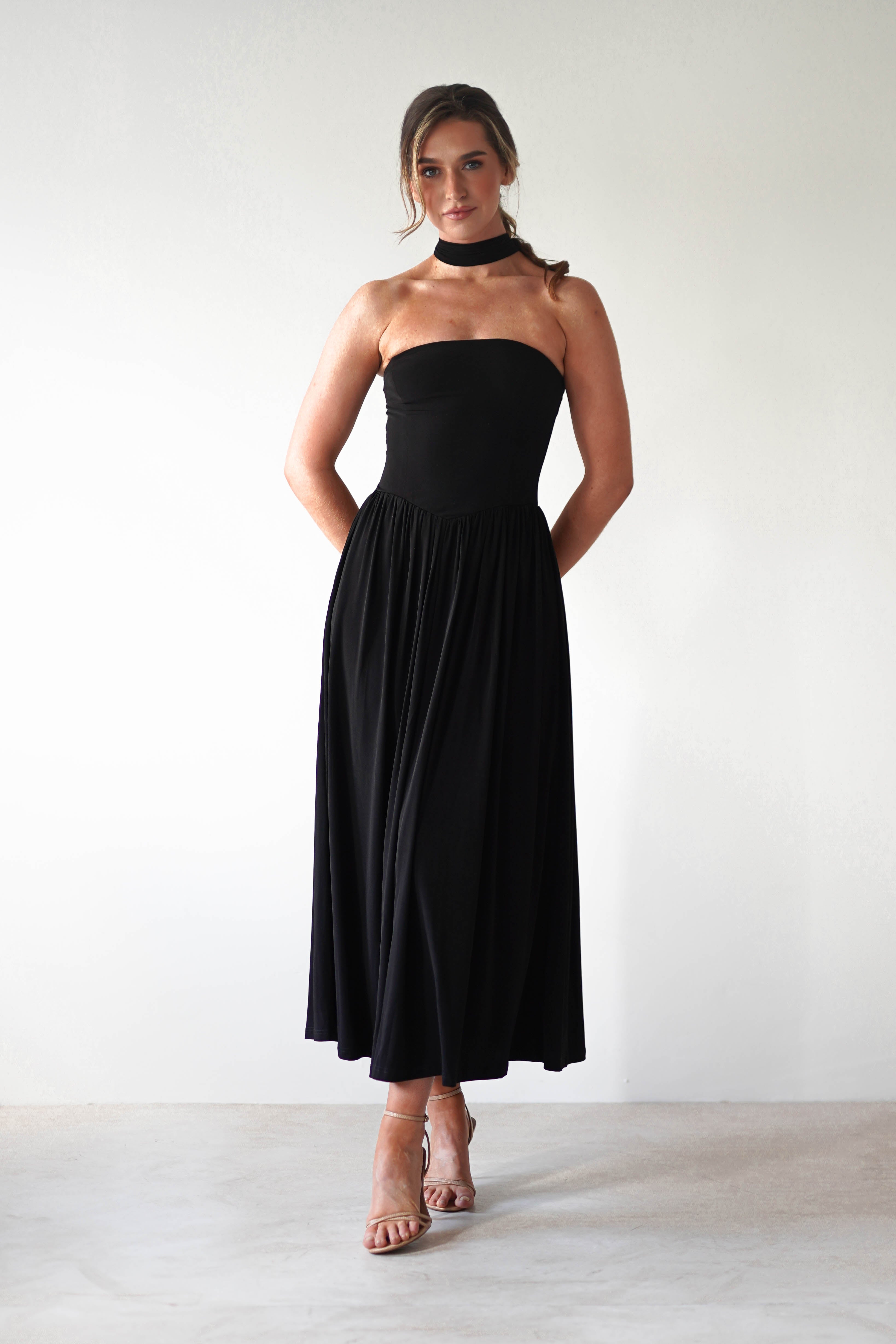 A woman stands against a plain light background wearing a strapless black midi dress with a fitted bodice, pleated skirt, and a matching black choker. She has her hands behind her back and is wearing strappy nude heels—perfect for a wedding guest look.