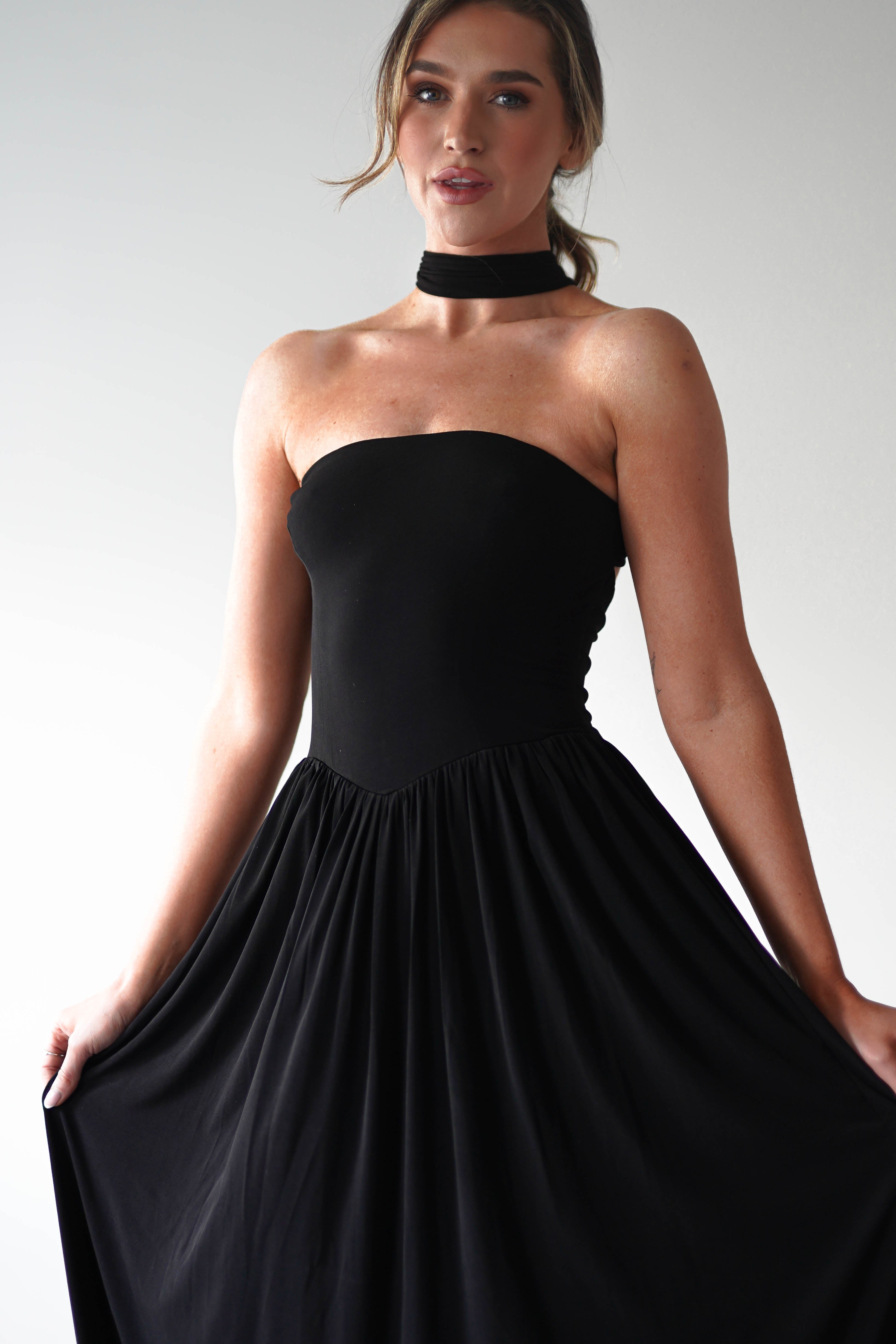 A woman wearing a strapless black dress with a pleated skirt and a matching black choker poses against a plain light background, holding the sides of her dress and looking slightly off-camera—an elegant look for any wedding guest.
