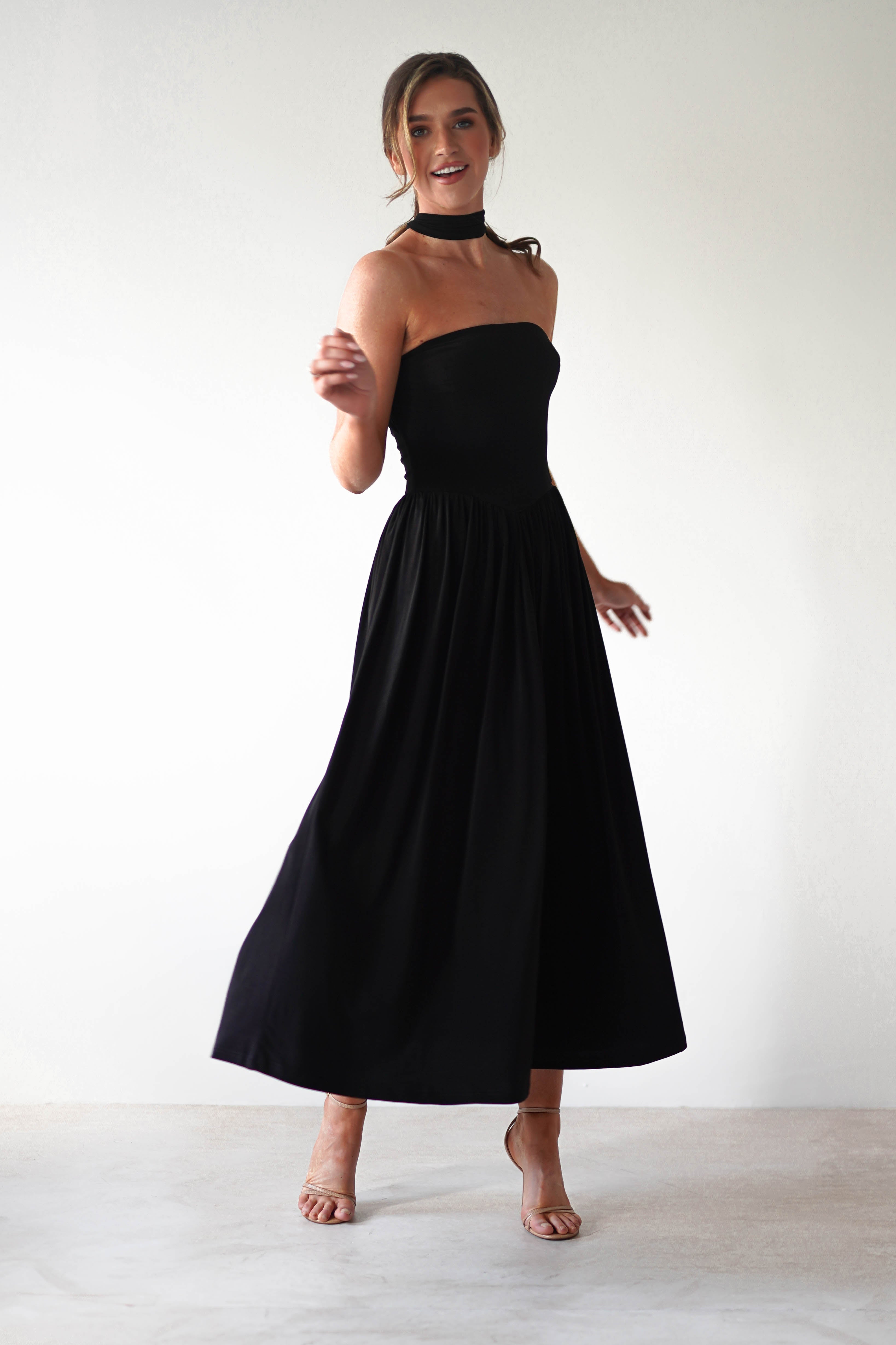 A woman wearing a strapless black midi dress with a fitted bodice and pleated skirt stands on light-colored flooring, posing with one arm bent and smiling. Perfect for a wedding guest look, she completes the outfit with a black choker and nude heeled sandals.