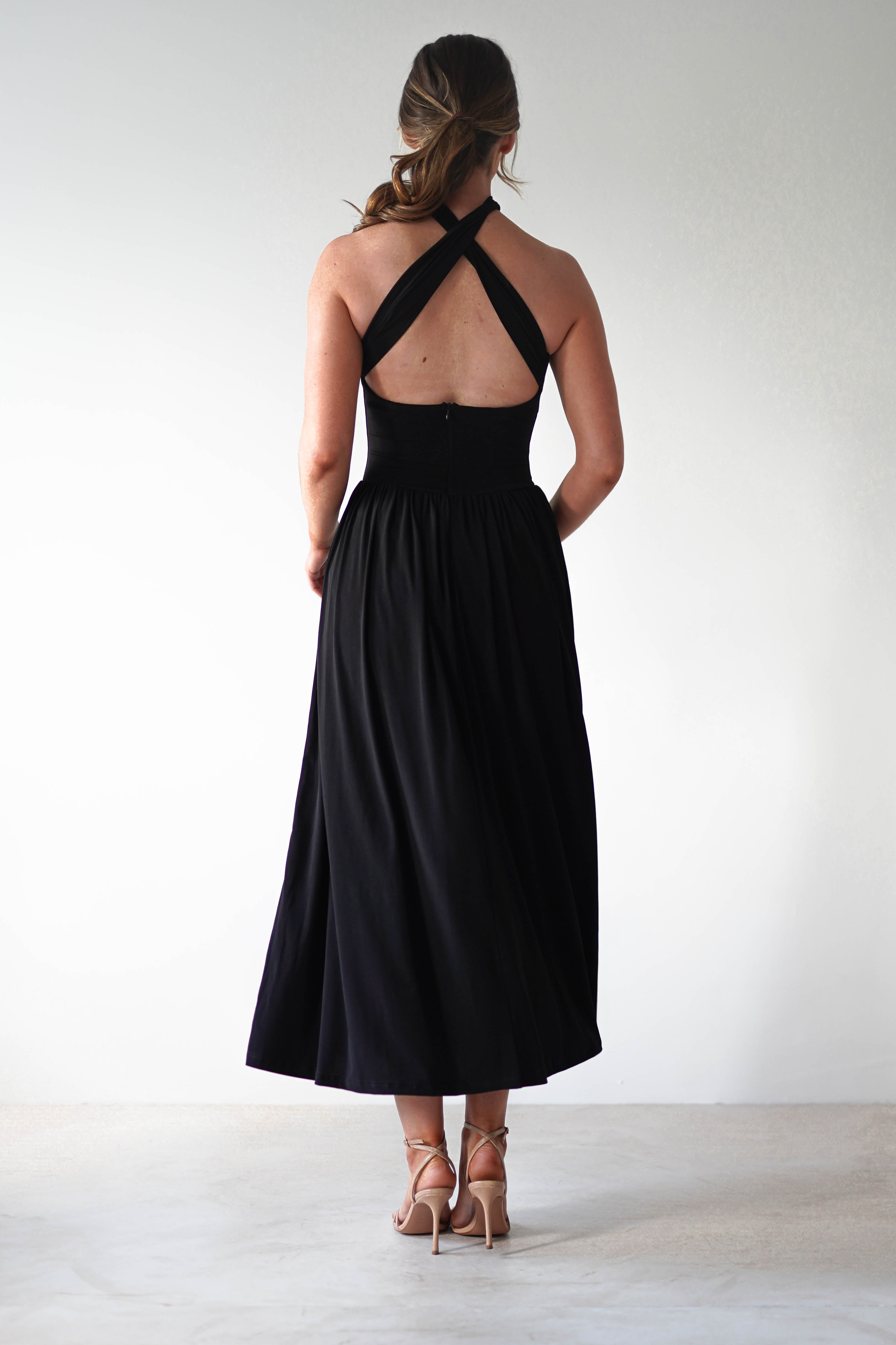 A woman stands facing away, wearing a sleeveless black midi dress with a crisscross halter back and pleated skirt, styled as the perfect wedding guest look with nude high-heeled sandals against a plain, light background.