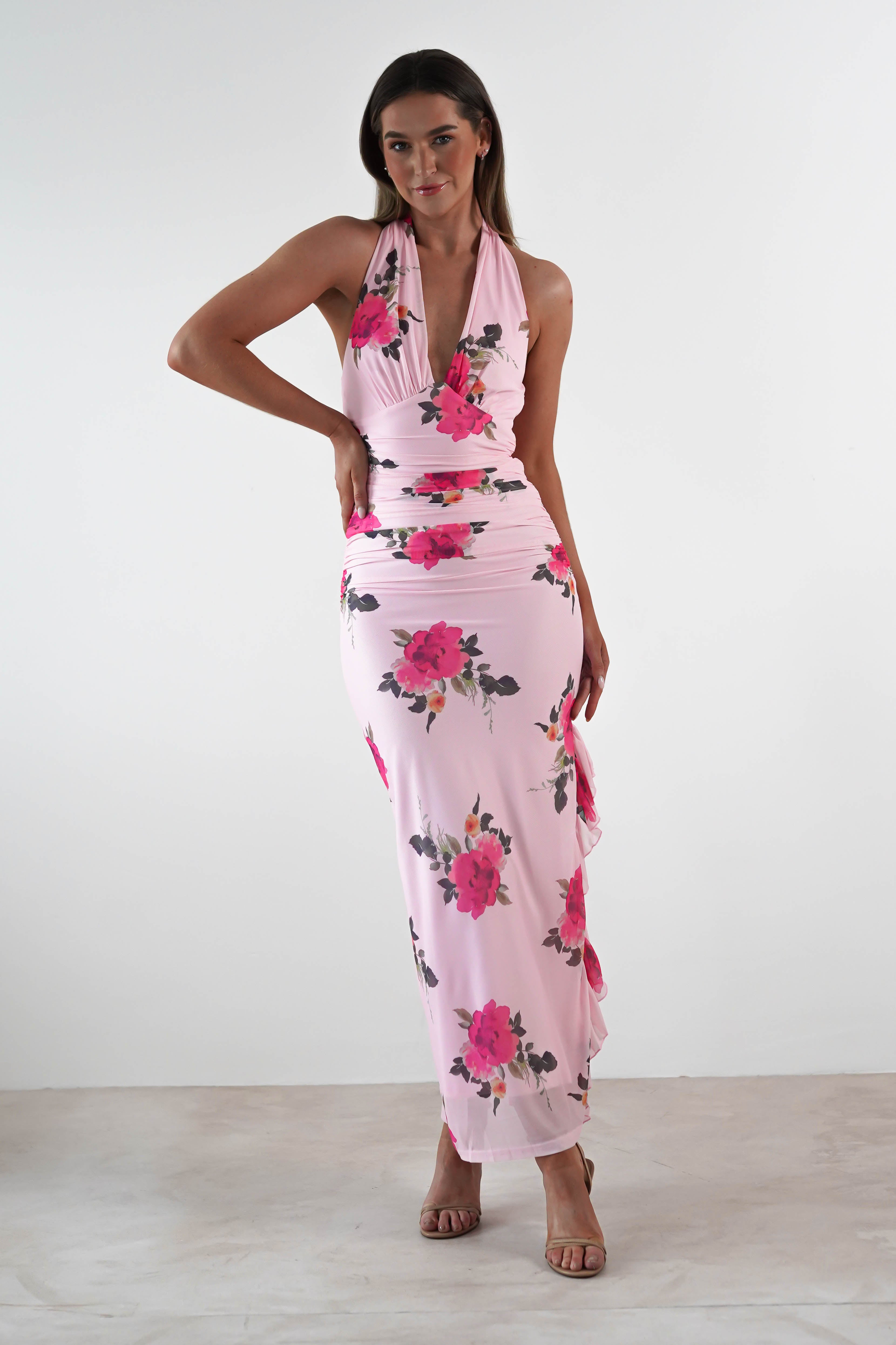 A woman poses against a plain white background wearing a sleeveless, floor-length maxi dress with a deep V-neck and bright pink floral print—an elegant choice for a wedding guest—paired with nude high-heeled sandals.