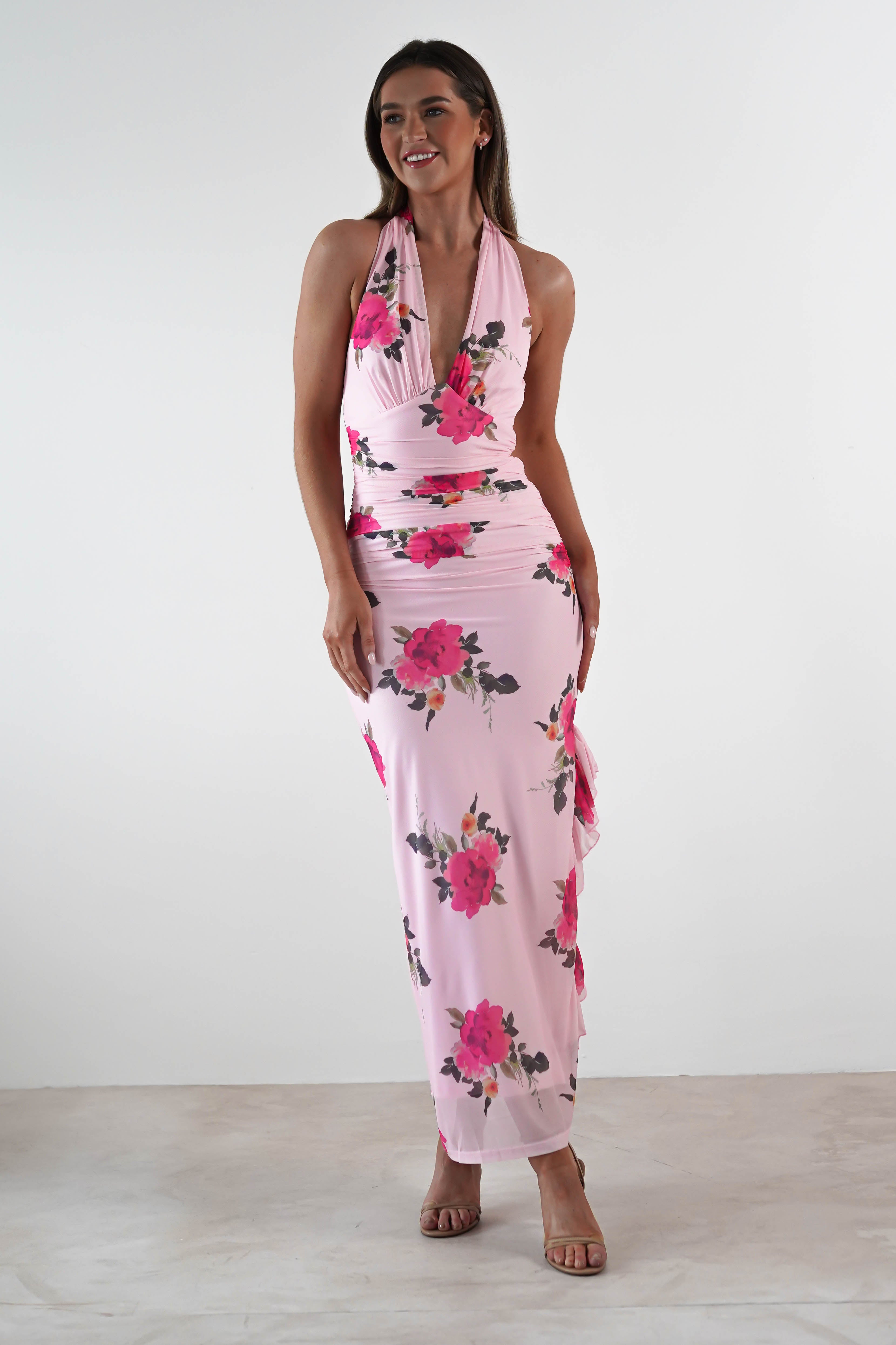 A woman wears a sleeveless, floor-length pink maxi dress with a halter neckline and bold red floral print, paired with nude heels—a perfect choice for a wedding guest—standing against a plain white background.