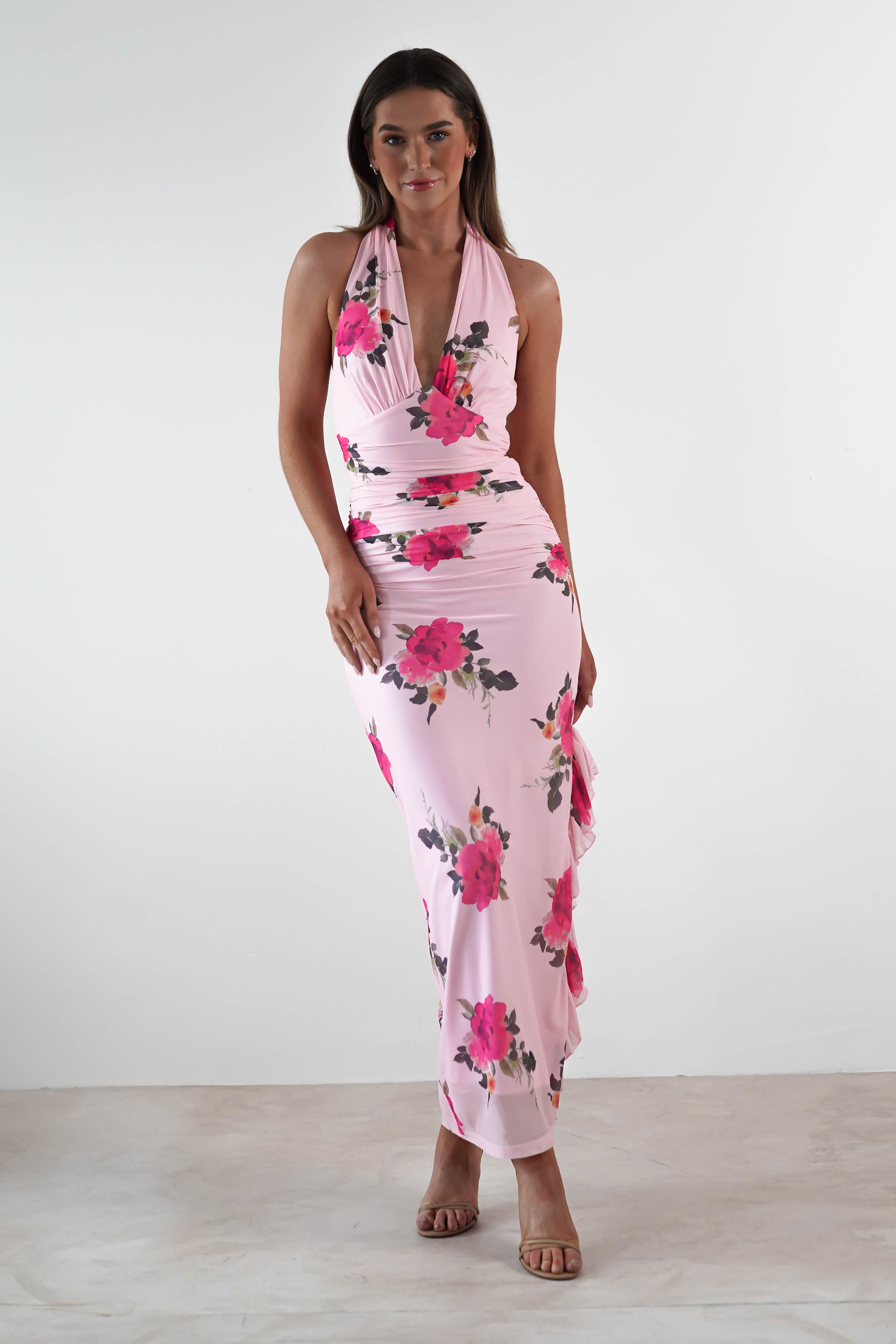 A woman stands against a plain white background wearing a sleeveless, light pink floral print maxi dress with ruching details, perfect for a wedding guest look, paired with nude strappy heels.
