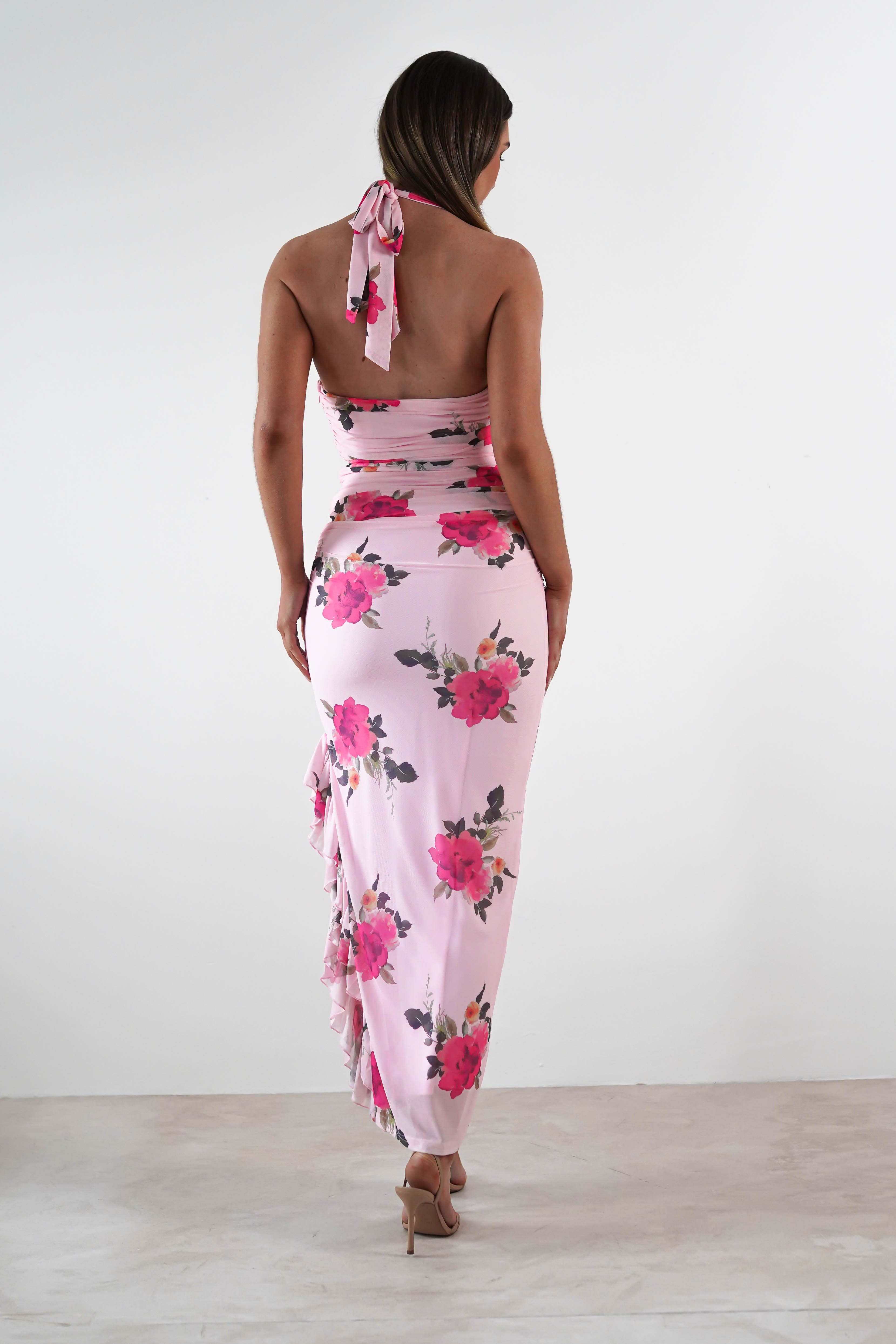 A woman stands facing away, wearing a light pink floral print maxi dress with an open back and tied halter straps—perfect for a wedding guest. She completes the look with nude high heels against a plain white background.