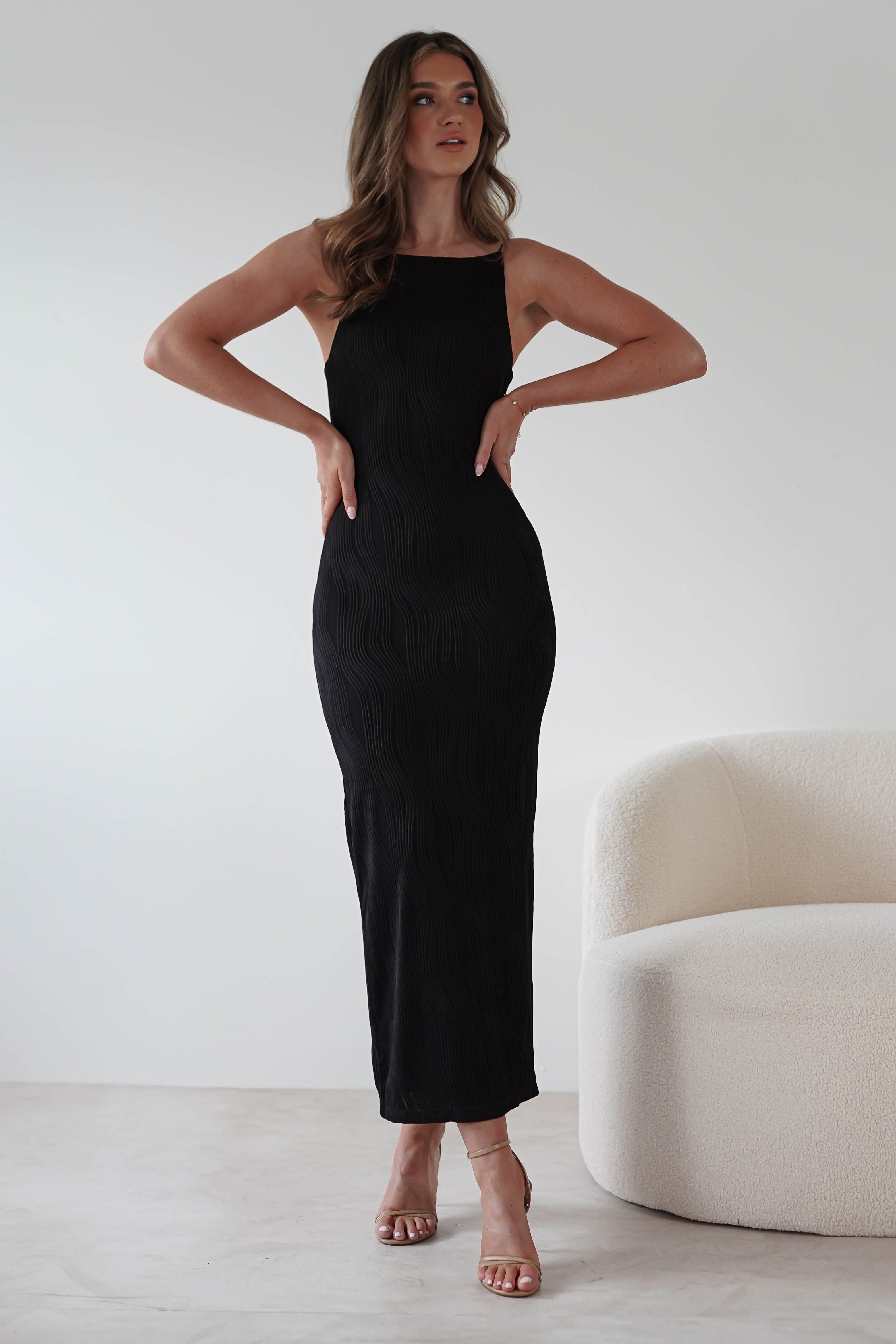 A woman stands in a minimalist room wearing a fitted, sleeveless textured midaxi dress and nude heels. Her hands are on her hips, and she has wavy hair. Perfect for special occasions, the scene is complemented by a curved white upholstered chair against a white wall.