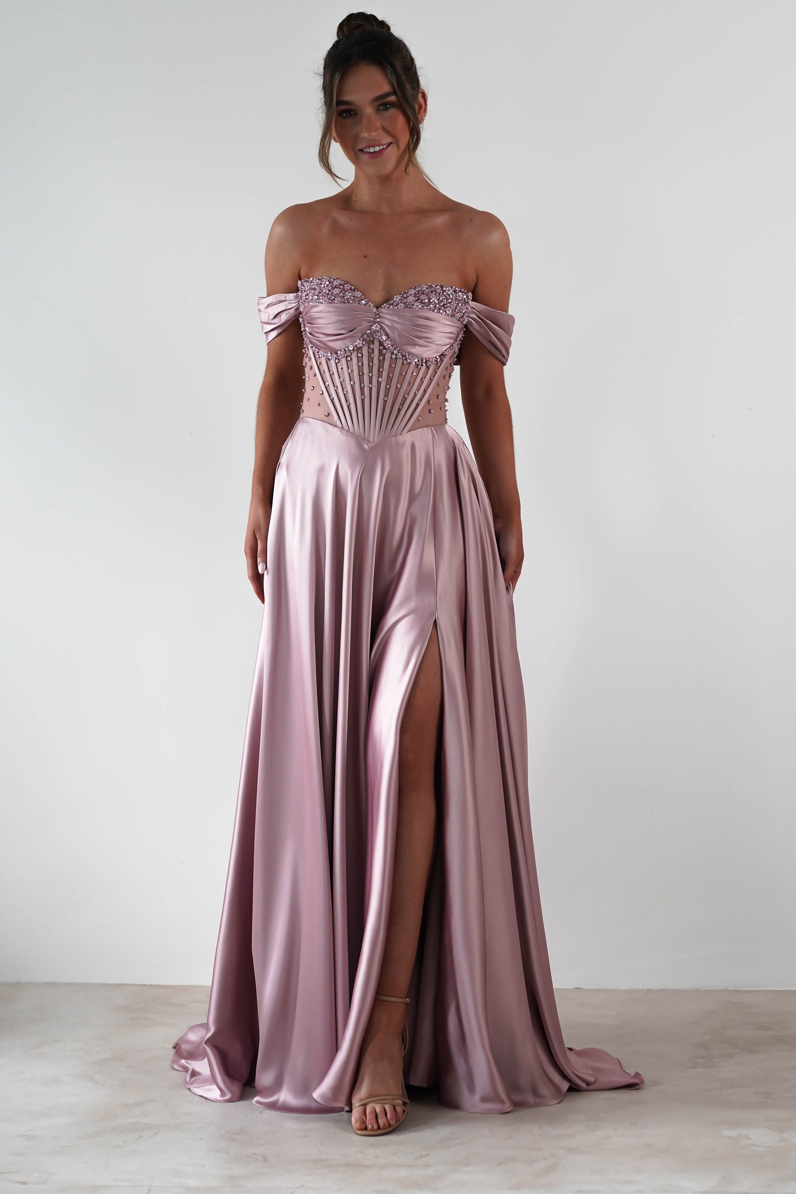 A woman wears a rose A-Line gown with a strapless sweetheart neckline, beaded bodice, and high slit, standing against a plain white background and smiling.