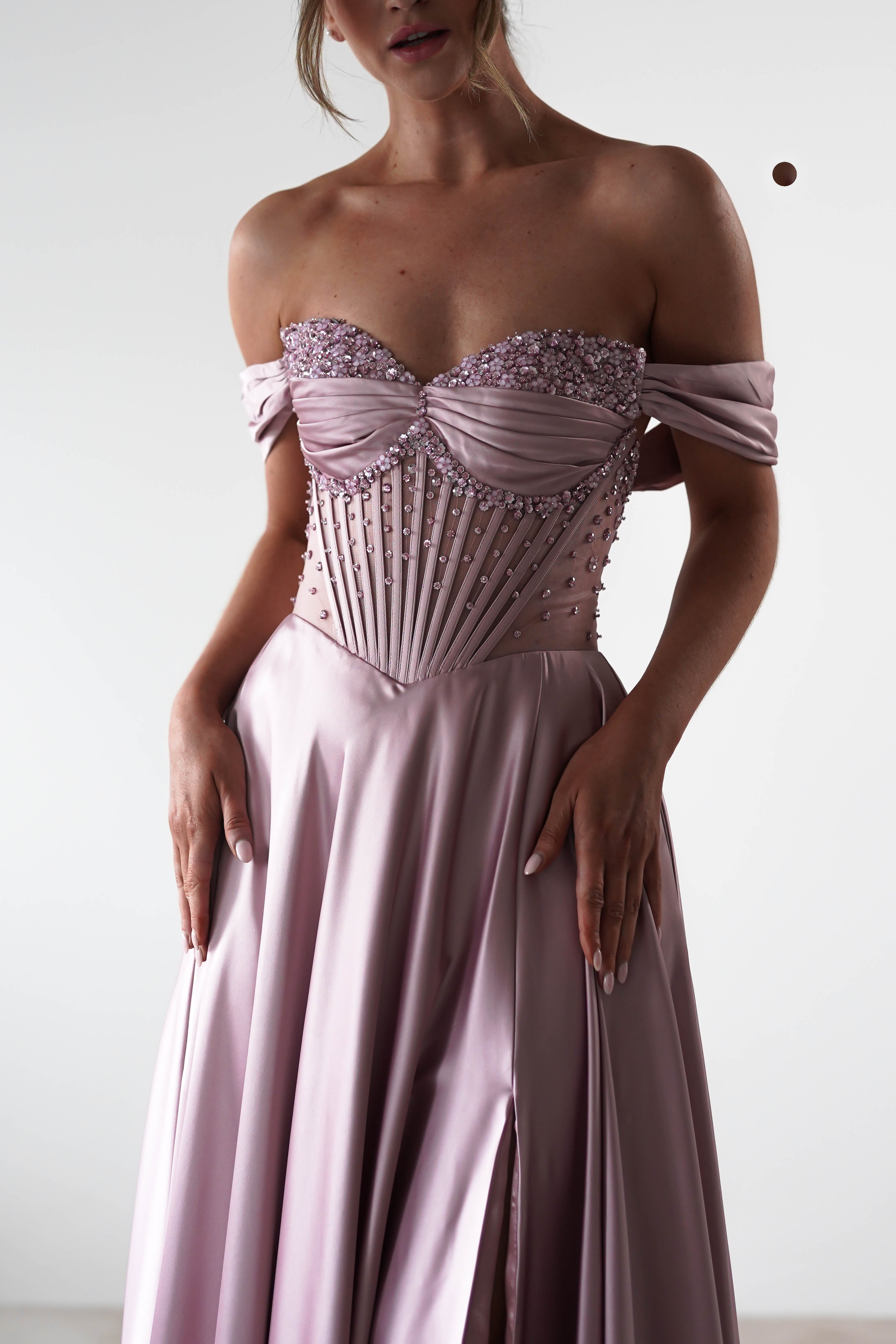 A woman wears a light purple rose A-Line gown with a beaded, structured bodice and flowing skirt. Her hands rest gently on the sides of the dress against a plain background.