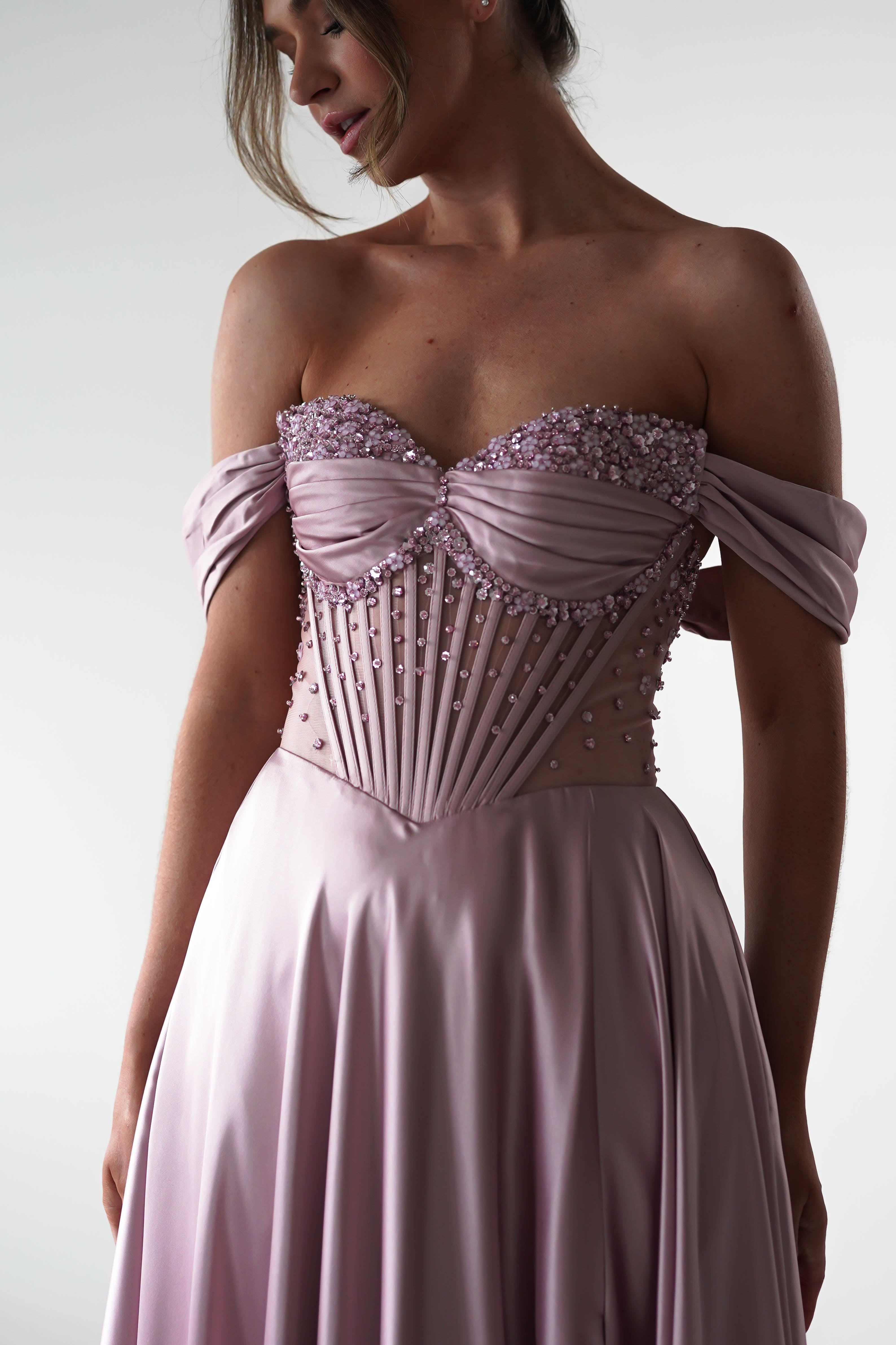 A woman in a strapless, rose A-Line gown with draped off-shoulder sleeves and a beaded, corset-style bodice adorned with pearls and pleats. Her head is slightly turned, hair swept up in an elegant updo.