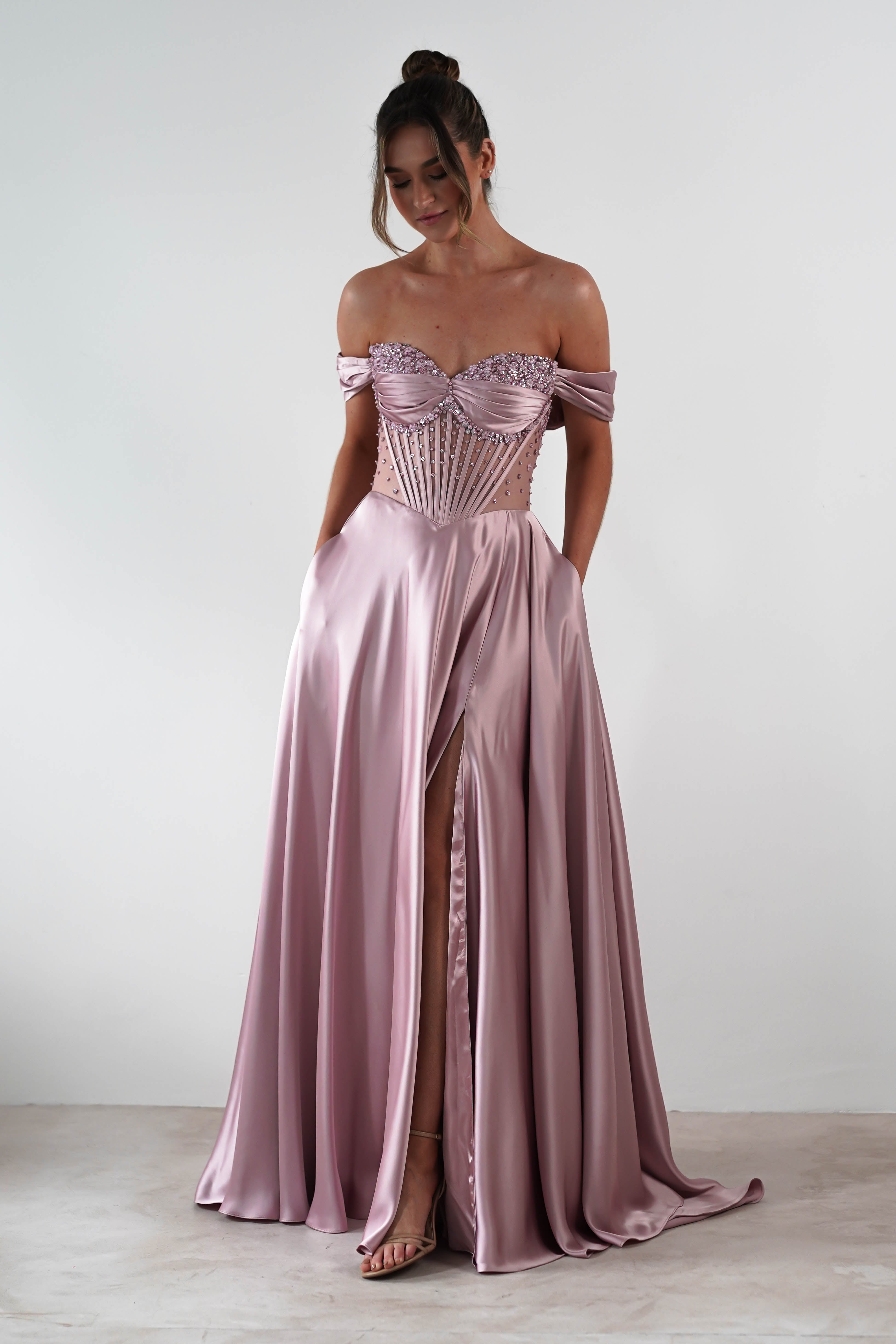 A woman in a rose A-Line gown with a strapless sweetheart neckline and floral embellishments on the structured bodice stands against a plain white background, looking down with her hands in the dress pockets.
