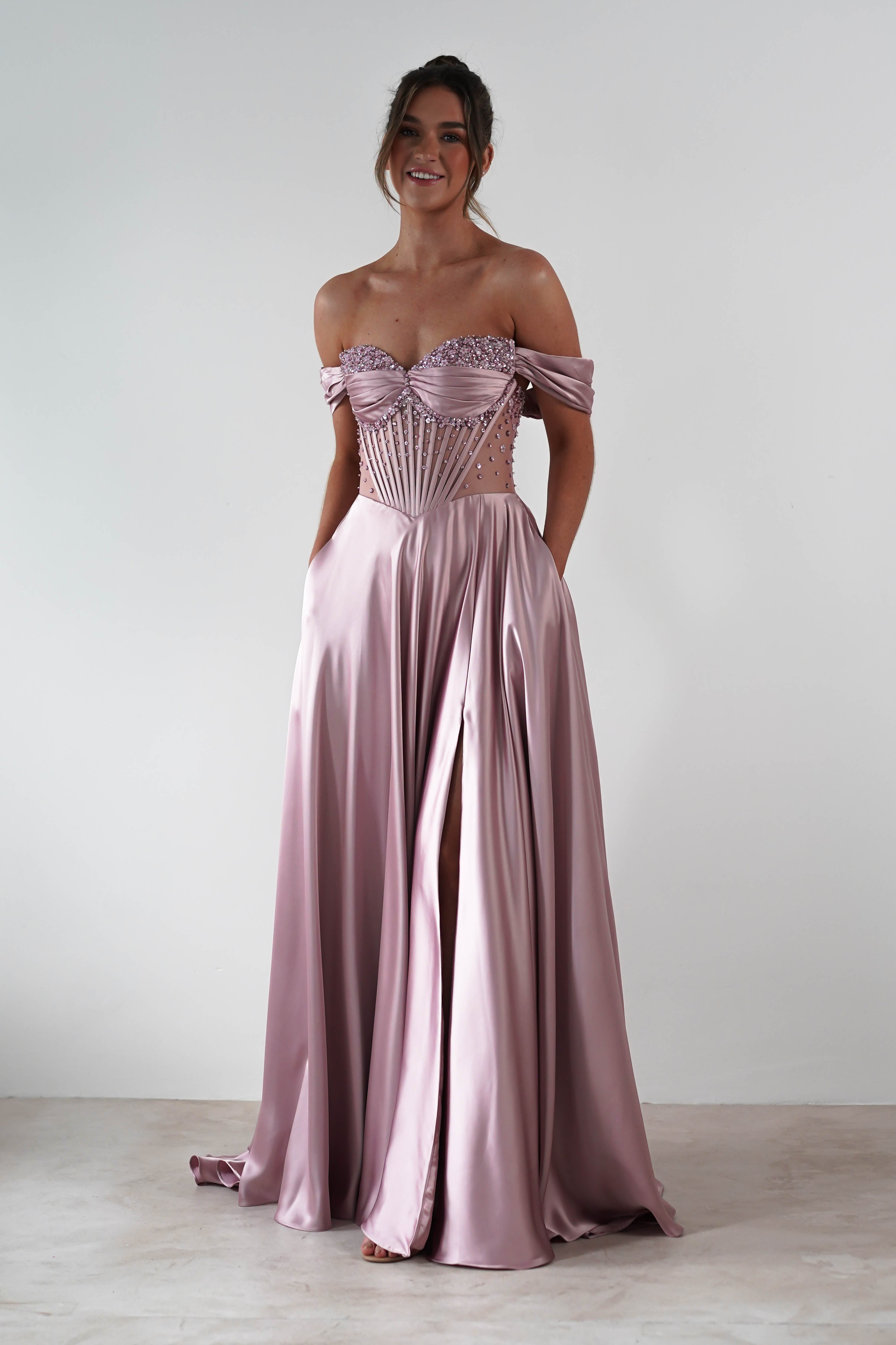 A woman stands against a plain background in a strapless sweetheart neckline, light mauve satin gown with a corset-style bodice, beaded details, and a high slit. Smiling, she slips her hands in her pockets.