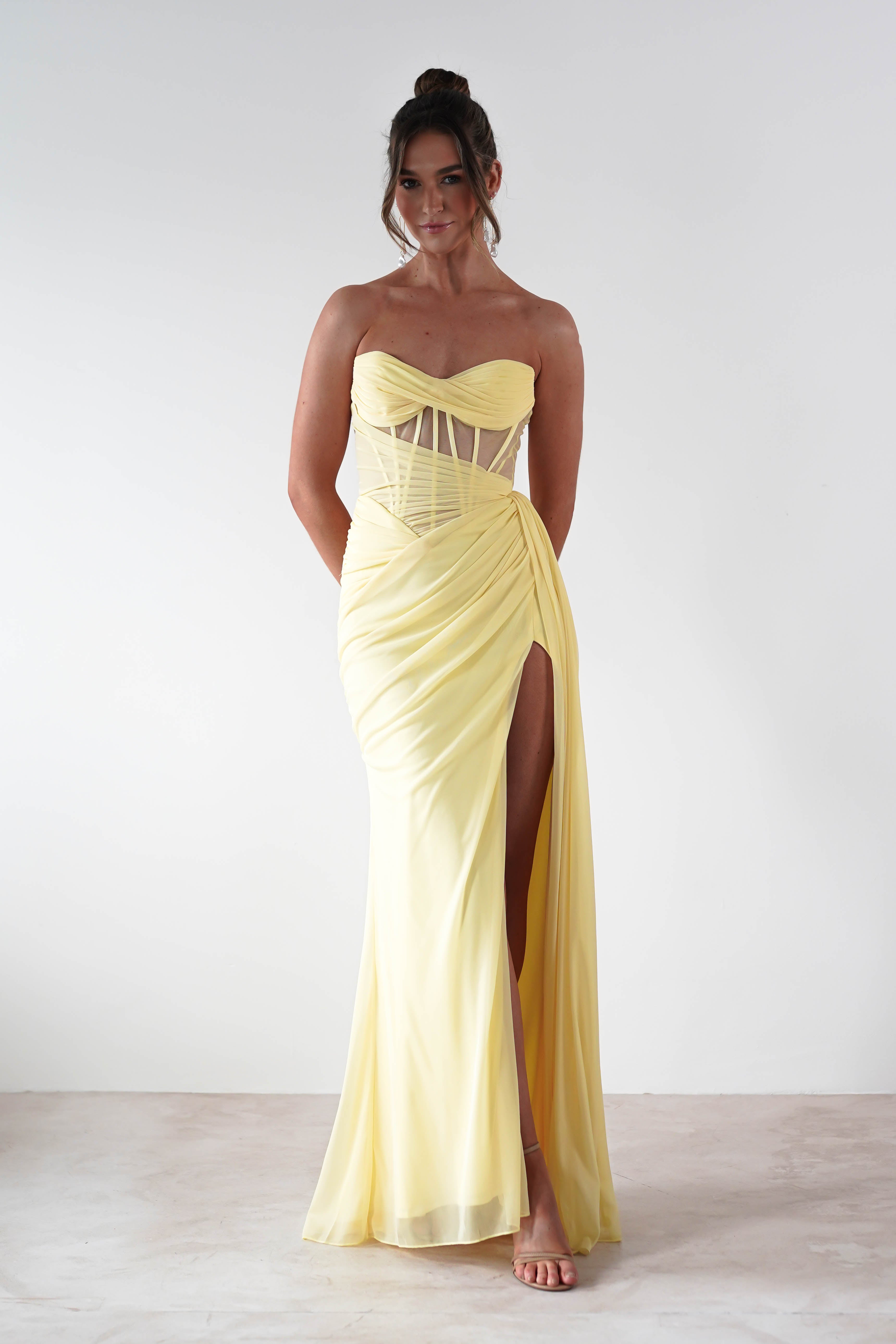A person poses in a floor-length, light yellow strapless evening gown featuring chiffon fabric and sheer details. The design includes a corset bodice and a thigh-high slit. The plain white background highlights the elegant dress.