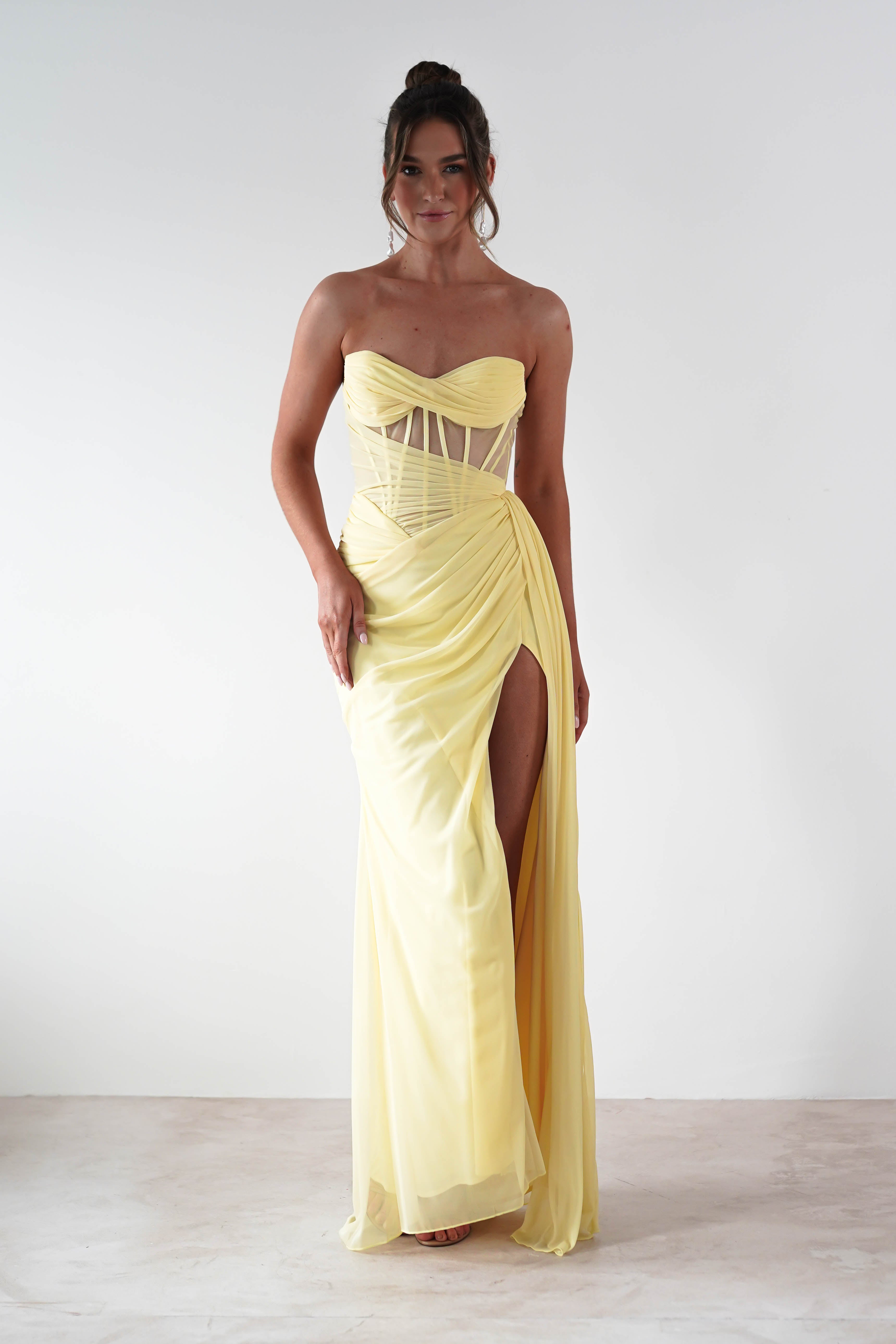 A woman stands against a plain background in a strapless evening gown, floor-length and yellow, featuring a corset-style bodice and high slit. The dress boasts a ruched design crafted from chiffon fabric, with her hair elegantly styled in an updo.