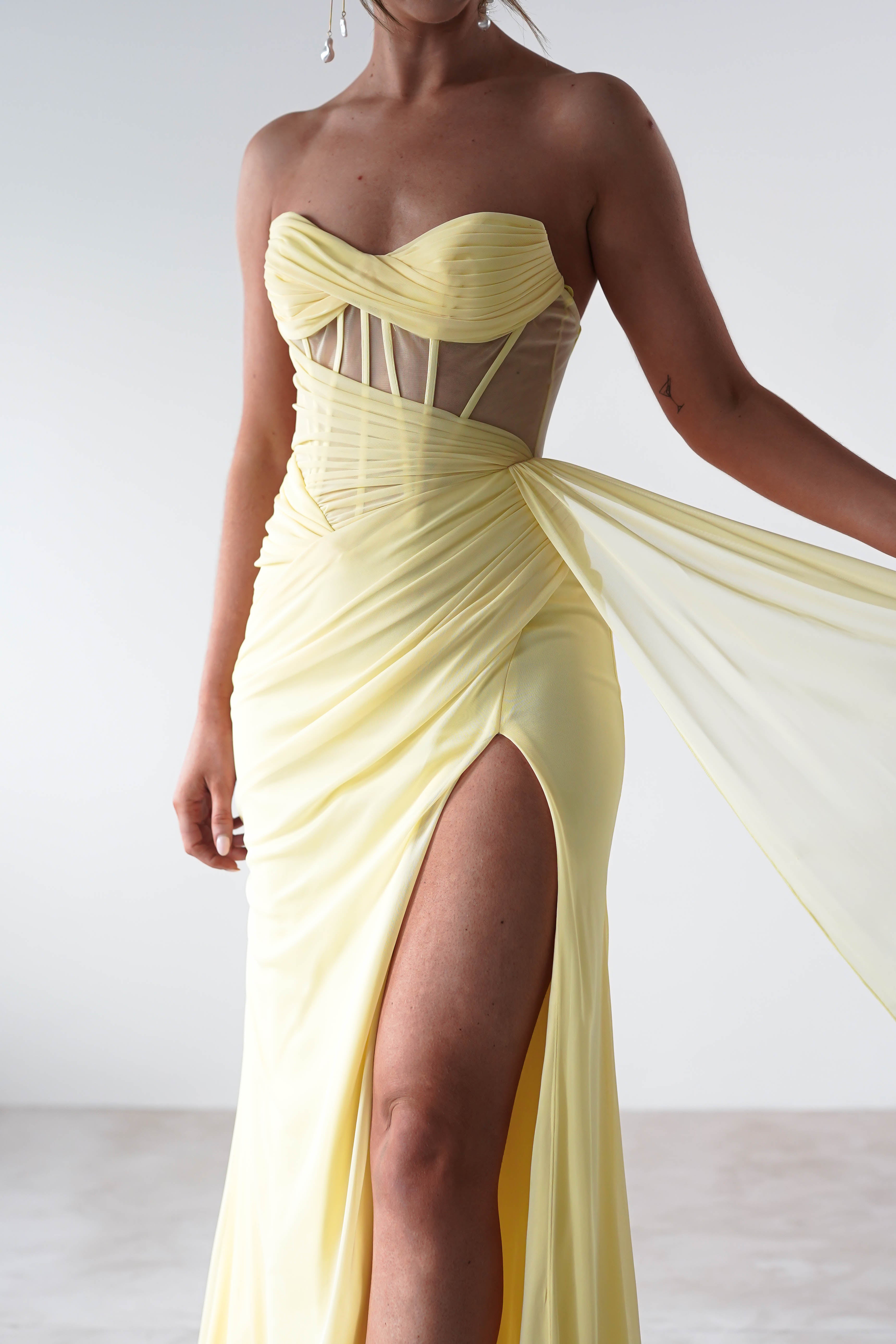 A person is wearing a strapless evening gown in pale yellow, featuring a high slit and sheer paneling. The fabric, possibly chiffon, is elegantly gathered at the waist as they hold a flowing piece attached to the dress. The background remains plain white.