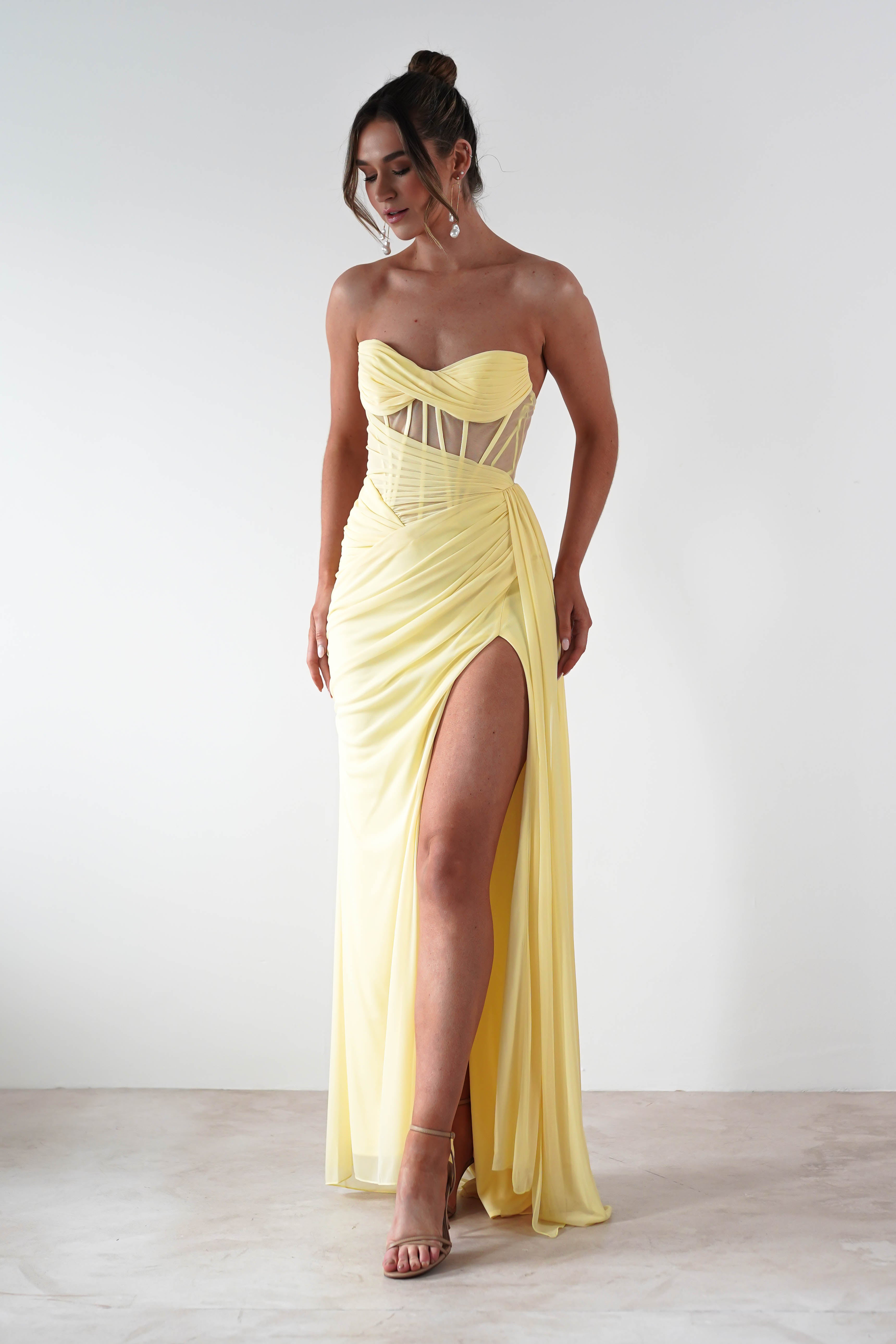 A woman wearing a strapless evening gown in pastel yellow chiffon fabric, featuring a corset bodice and a high slit, stands against a plain background. Her hair is in an elegant updo and shes wearing high-heeled sandals.
