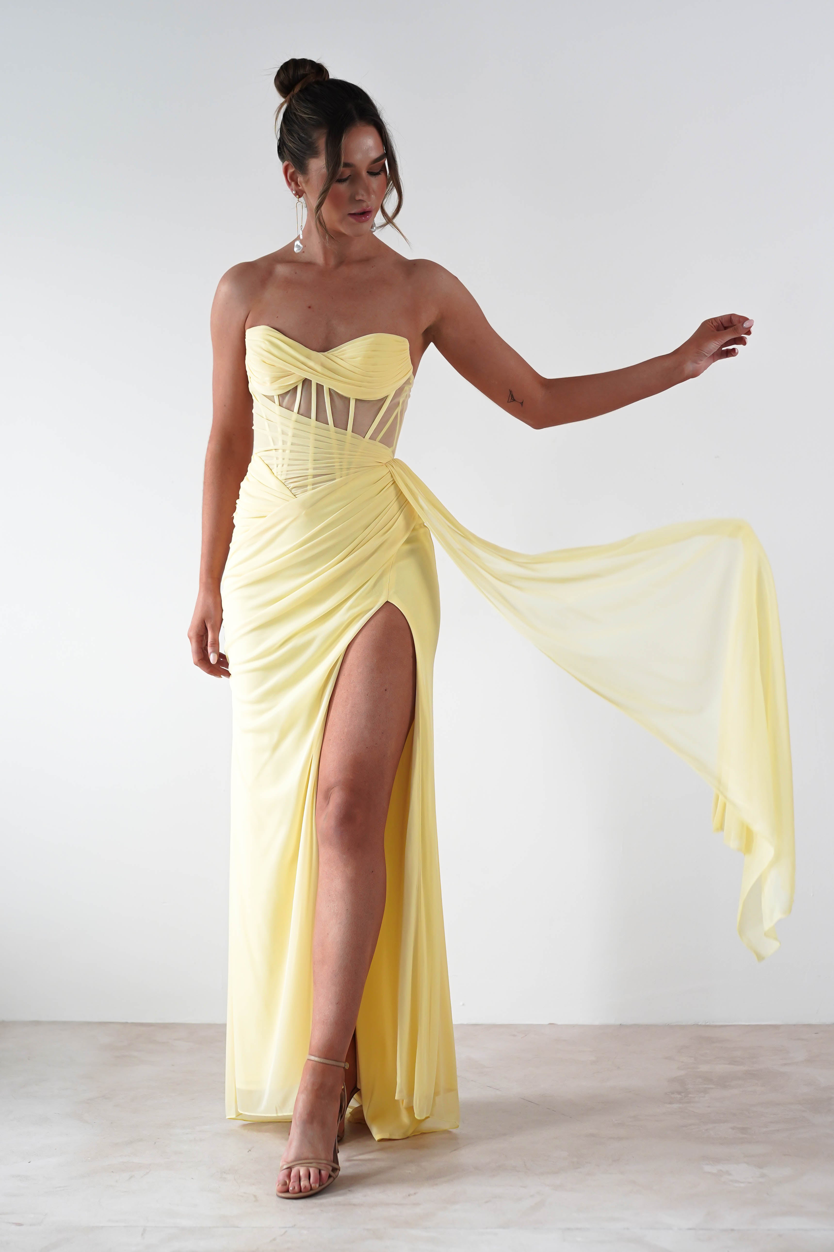 A woman in a strapless evening gown, featuring a sheer corset bodice and high slit, poses gracefully against a plain backdrop. With her hair styled in a bun and wearing heels, she elegantly holds the flowing chiffon fabric of her light yellow dress.
