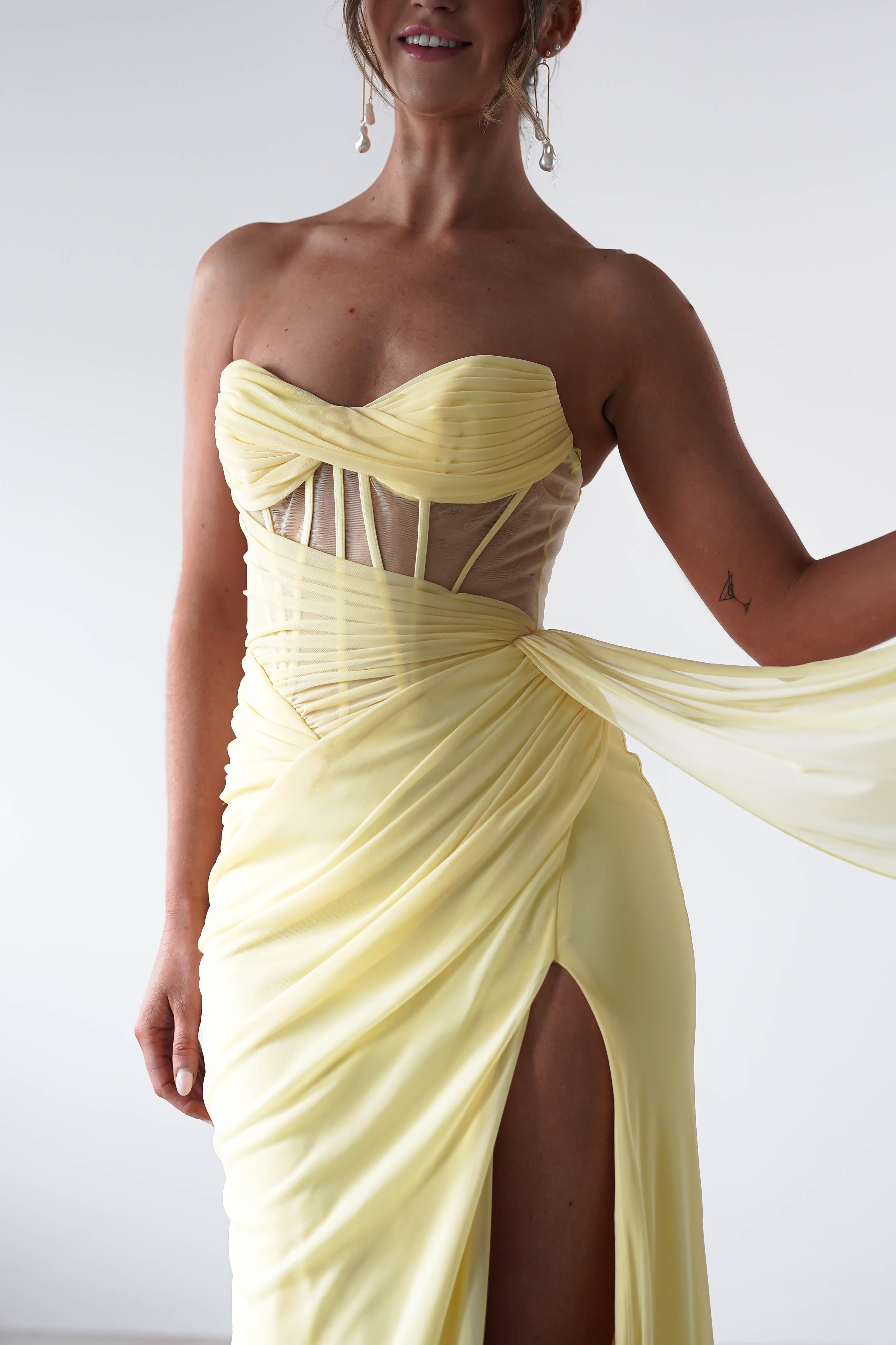 A person wearing a strapless evening gown in light yellow stands against a plain background. The gown, with its sweetheart neckline and structured corset bodice, features draped chiffon fabric and a high slit. The person elegantly holds part of the flowing drape.