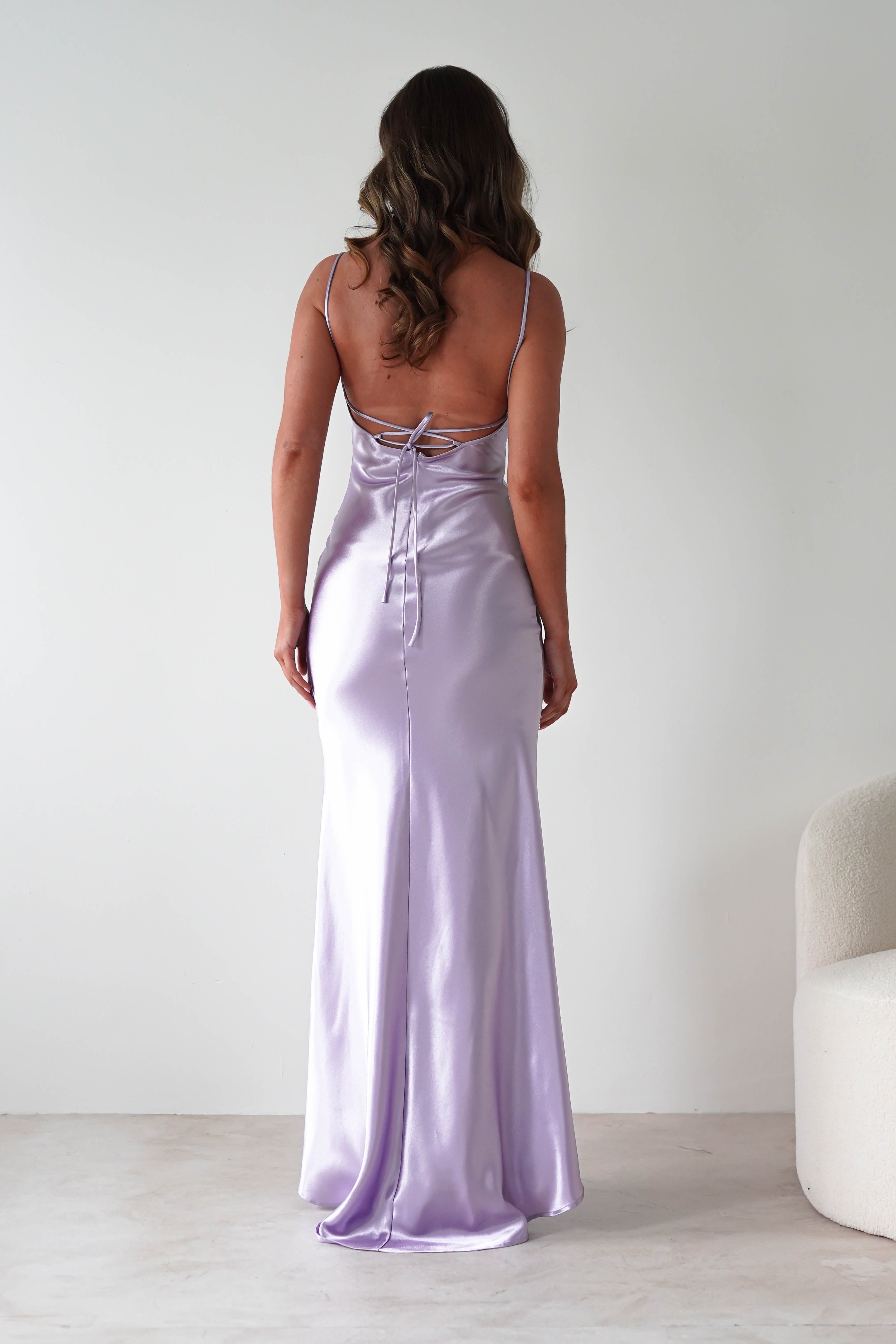 A woman with long, wavy hair stands facing away in a body-skimming silhouette, wearing a shiny lavender satin gown with thin straps and an open back. The minimal room features a white chair nearby.