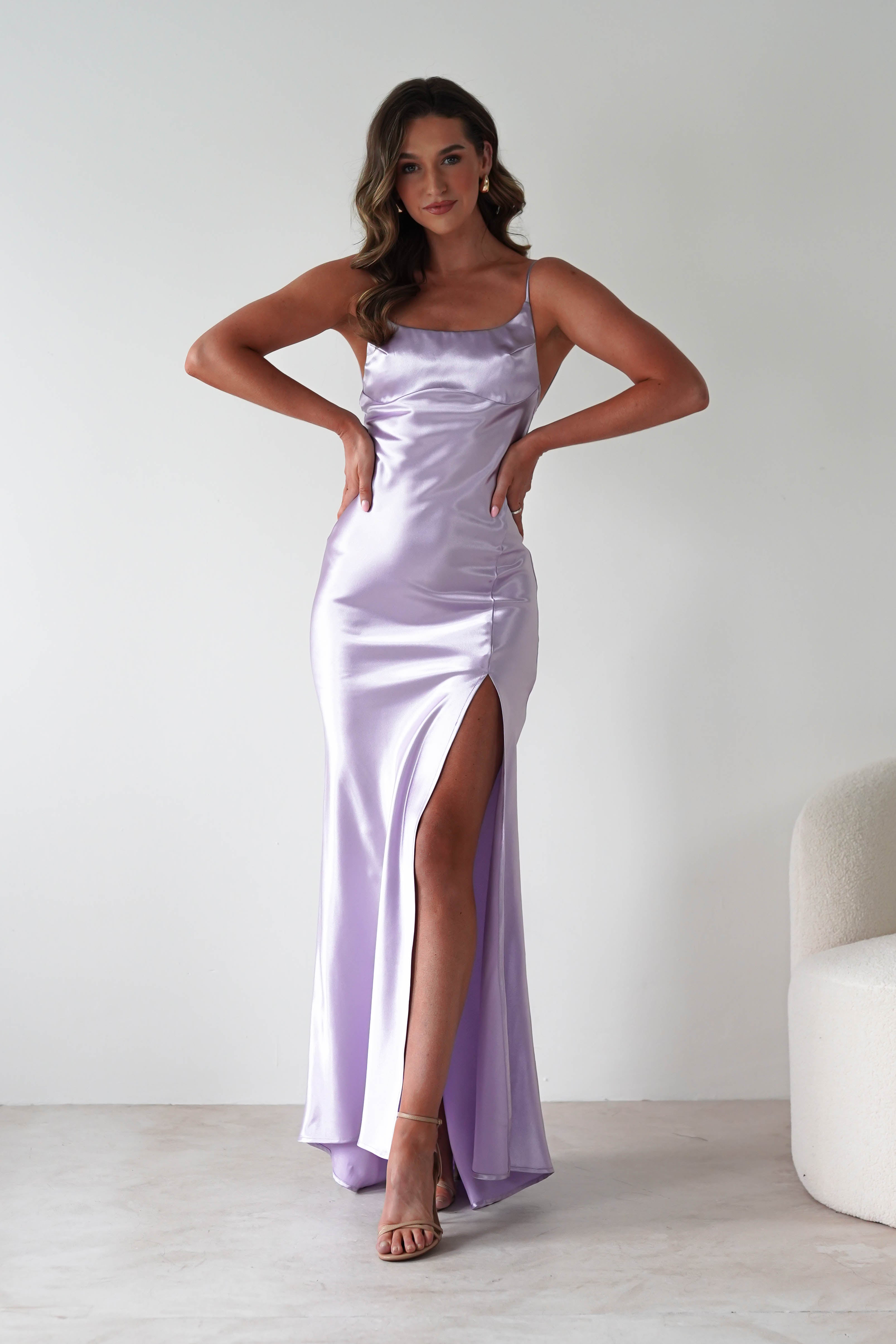 A woman stands in a lilac dress with a body-skimming silhouette, thin straps, a thigh-high slit, and neutral heels, posing with her hands on her hips against a plain white background next to a cream-colored chair.