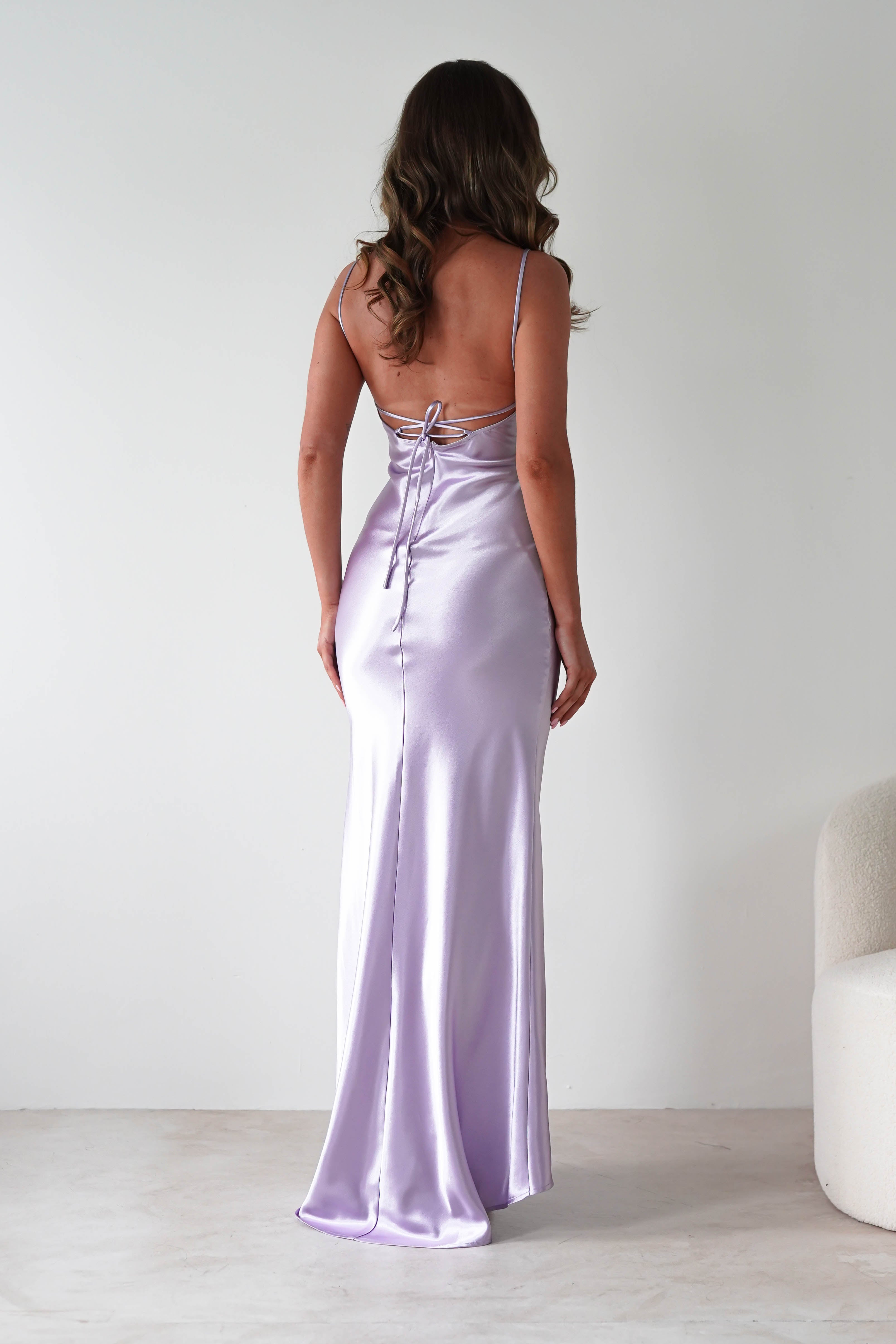 A woman with wavy brown hair wears a long, lilac satin dress with thin straps and an open back. The body-skimming silhouette highlights her figure as she stands indoors, facing away from the camera.