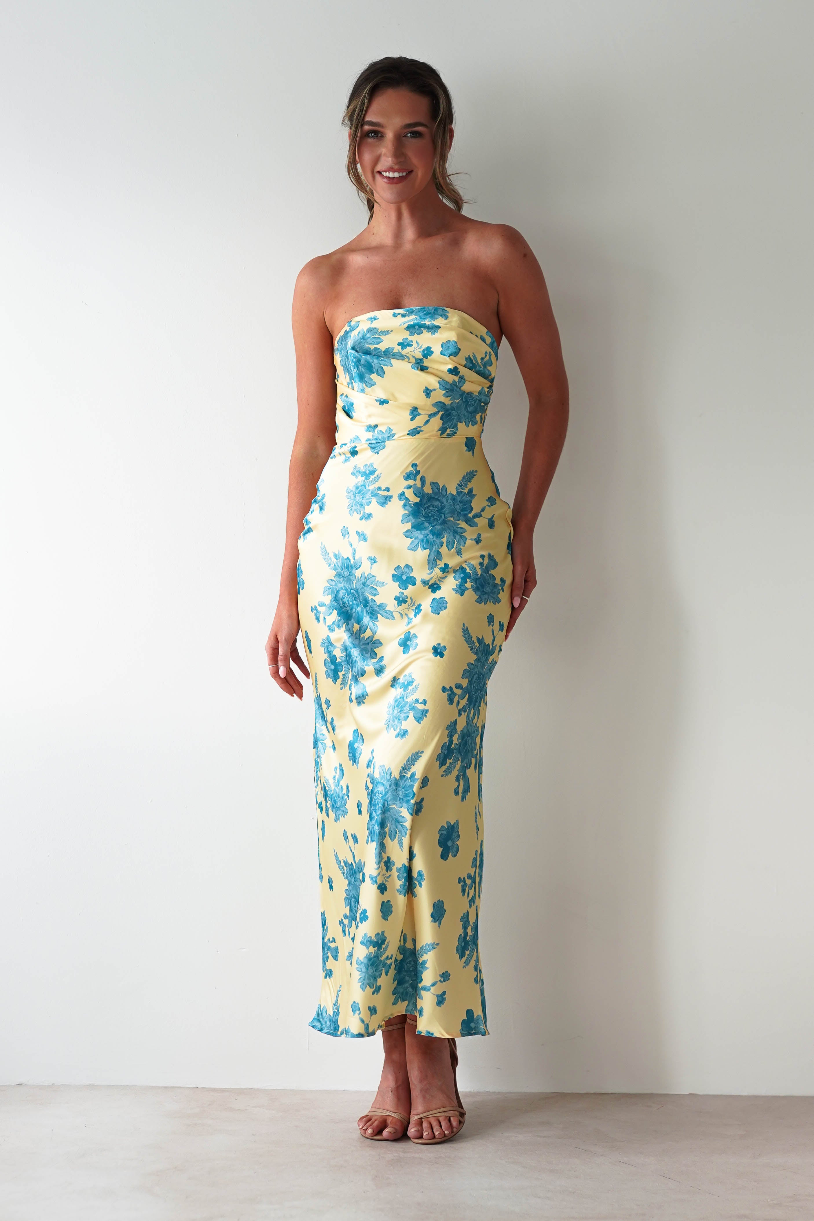 A woman stands against a white wall wearing a strapless satin maxi dress in yellow with blue floral patterns and nude high-heeled sandals. She is smiling with her hair tied back, making this the perfect wedding guest dress.