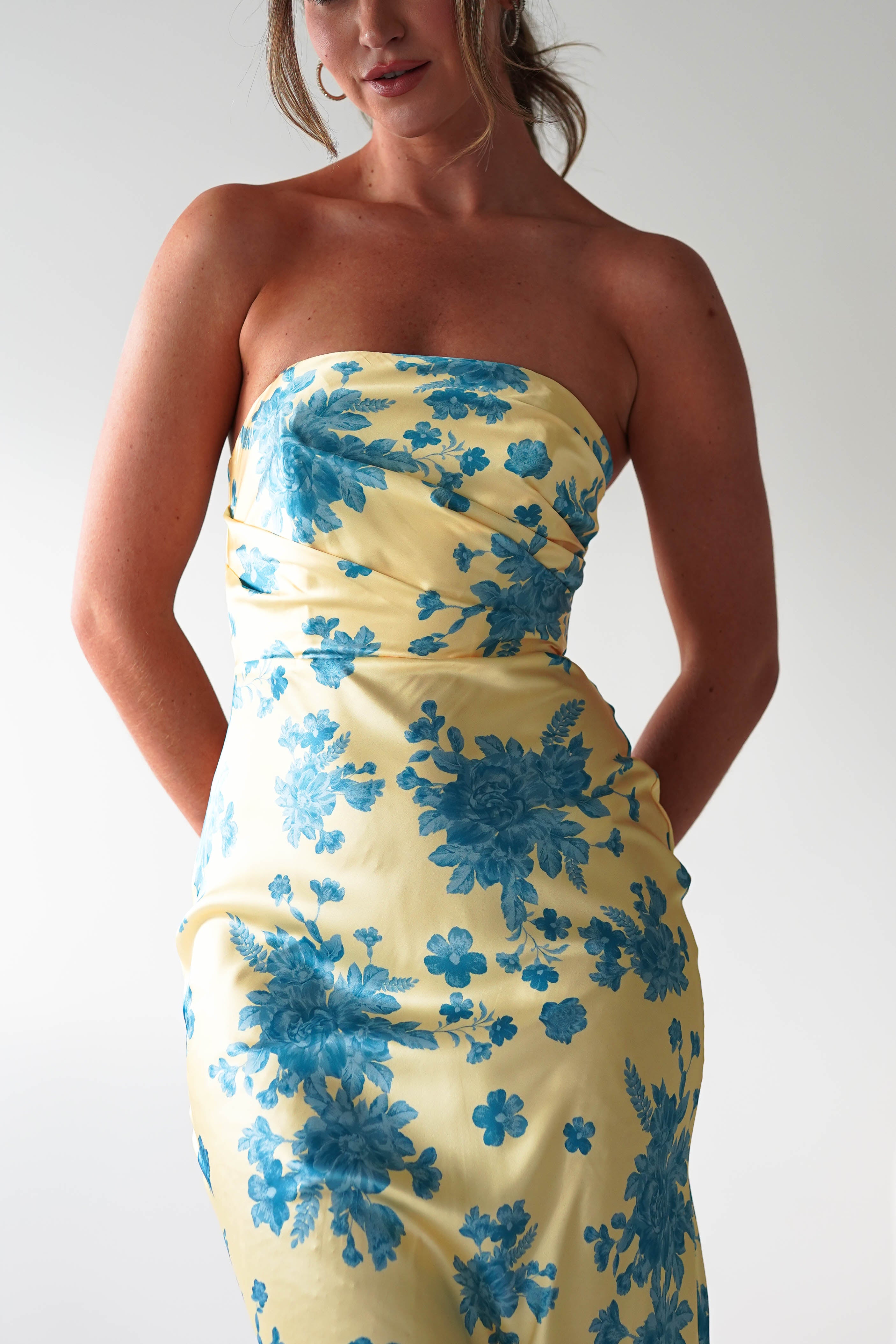 A woman wearing a strapless satin maxi dress in yellow with a blue floral pattern stands against a plain light background, her arms relaxed at her sides—an elegant choice for a wedding guest dress.
