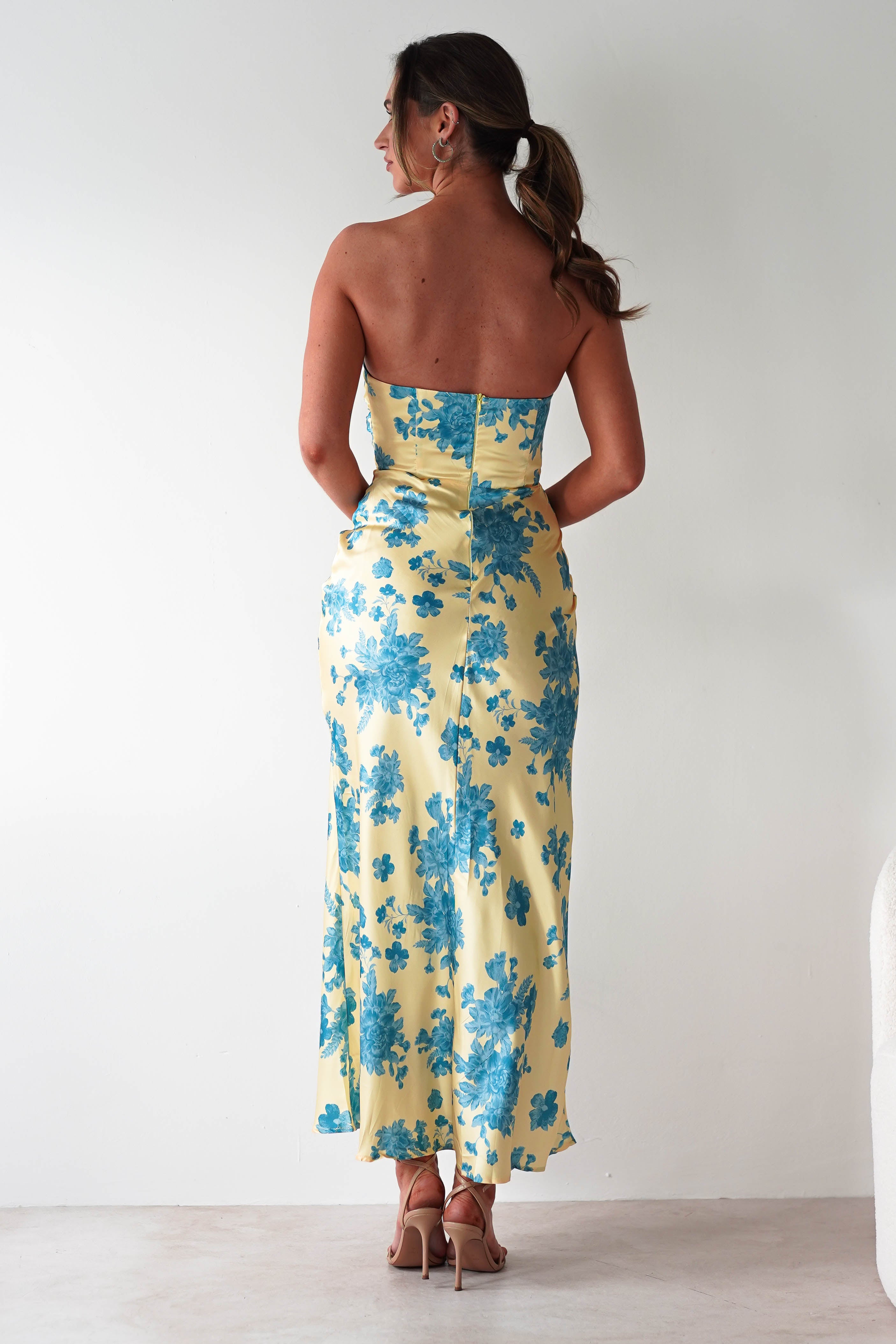 A woman with brown hair in a ponytail stands facing away, wearing a yellow strapless satin maxi dress with blue floral patterns and nude high-heeled sandals, perfect as a wedding guest dress, against a plain white background.