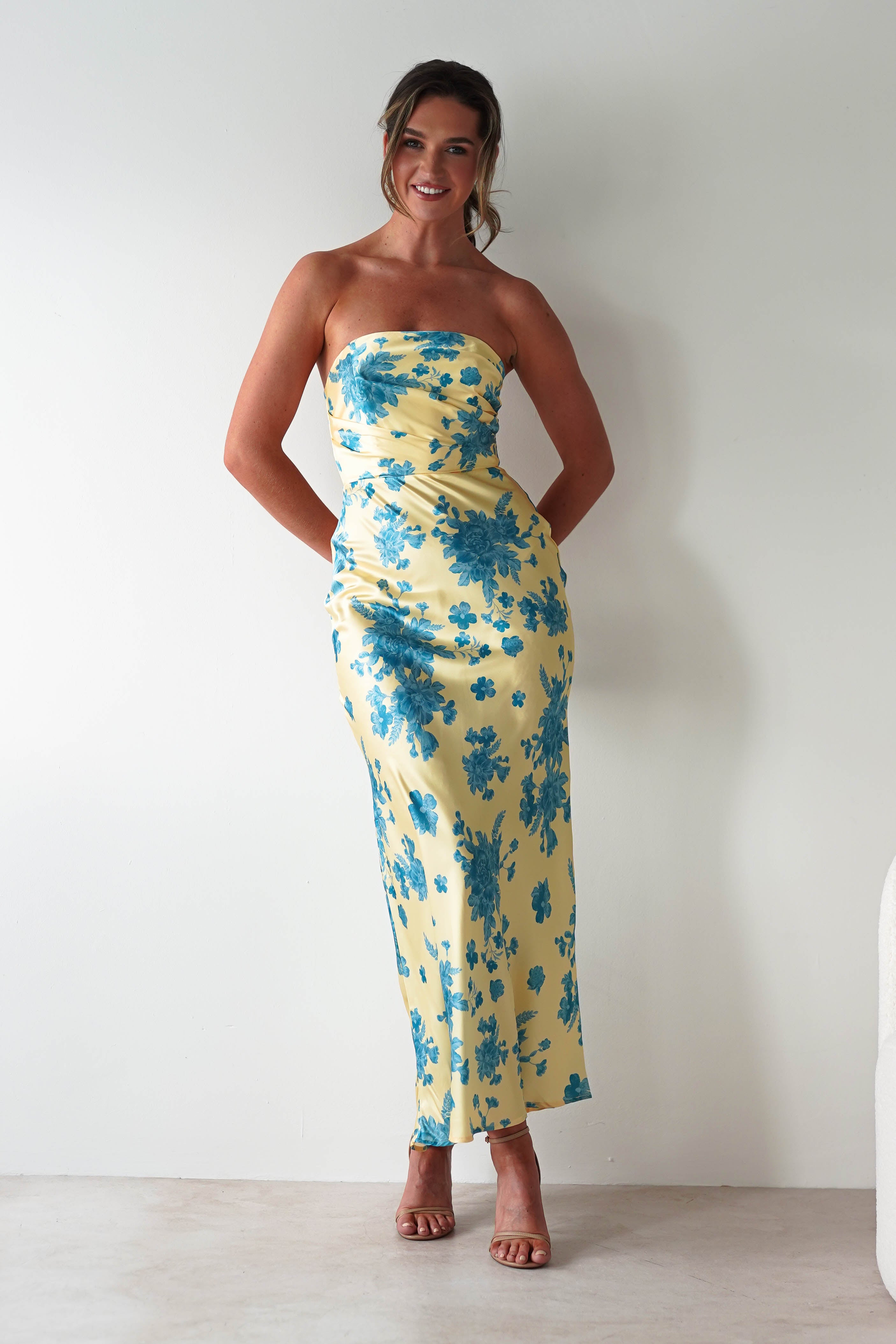 A woman stands smiling against a white wall, wearing a strapless satin maxi dress in yellow with blue floral patterns and brown heeled sandals—an effortlessly chic look perfect as a wedding guest dress. Her hair is styled up, hands tucked in the dress pockets.
