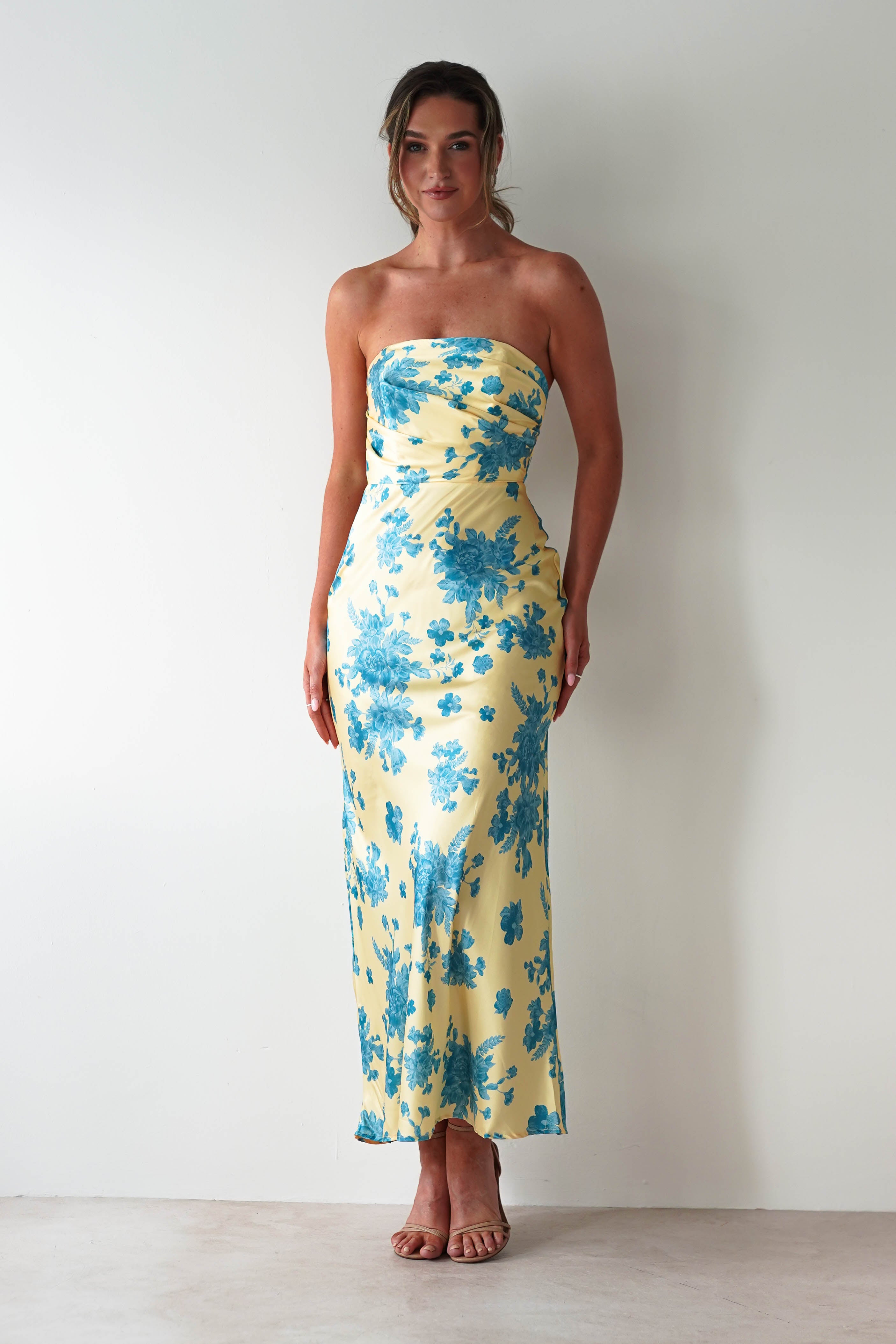 A woman stands against a white wall wearing a strapless satin maxi dress in yellow with blue floral patterns. She has her hair pulled back and is wearing nude heels, making this the perfect wedding guest dress.